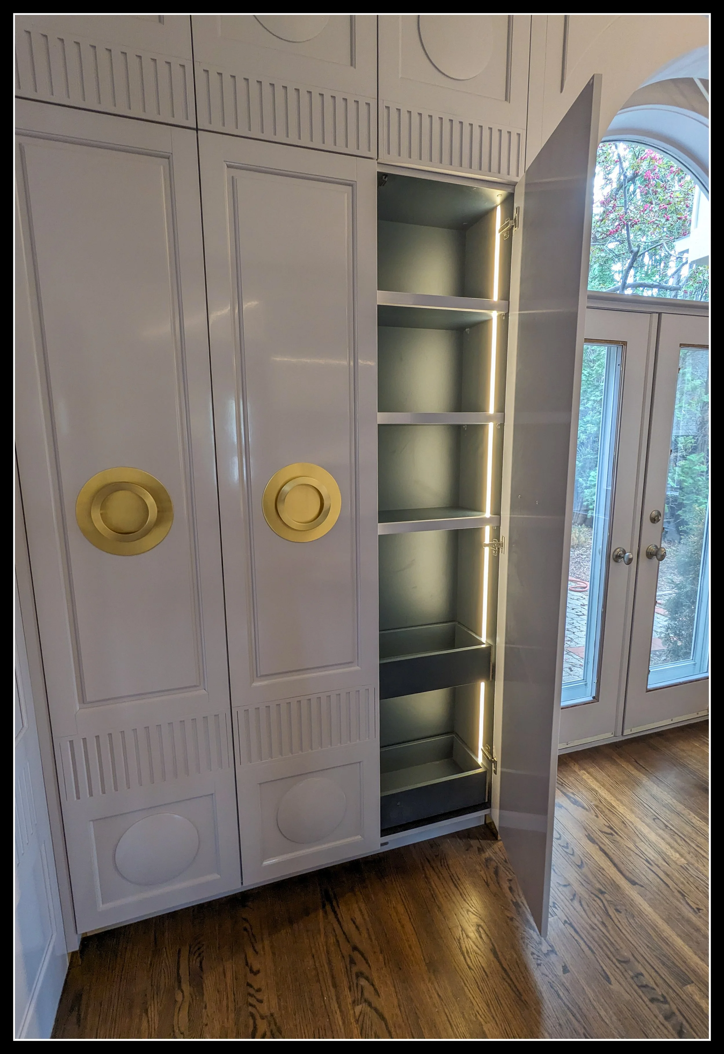 Tall gloss‑grey cabinet with fluted doors, lathe‑turned raised panels, solid brass pulls, and integrated vertical LED lighting in a premier architectural room.