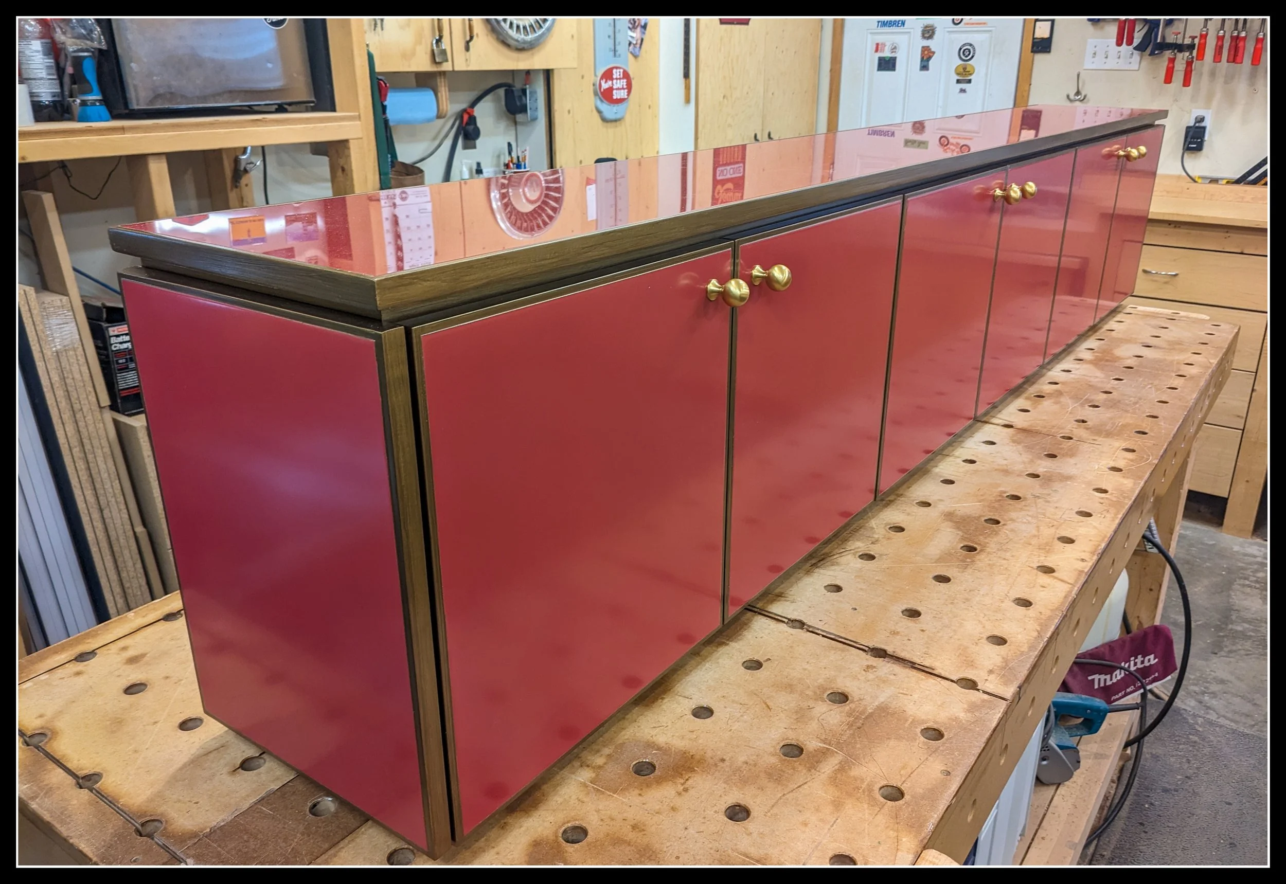 Gloss‑red cabinet with matching painted‑glass back panel, brass knobs, and walnut accents on a workbench in a woodworking shop.