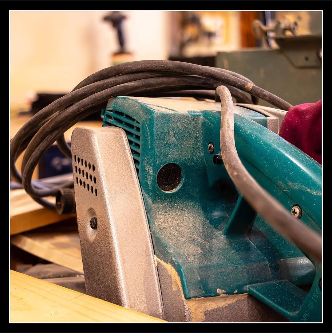 Makita 4-inch belt sander on a workbench with dust on the housing and cord wrapped around the tool in a woodworking shop.