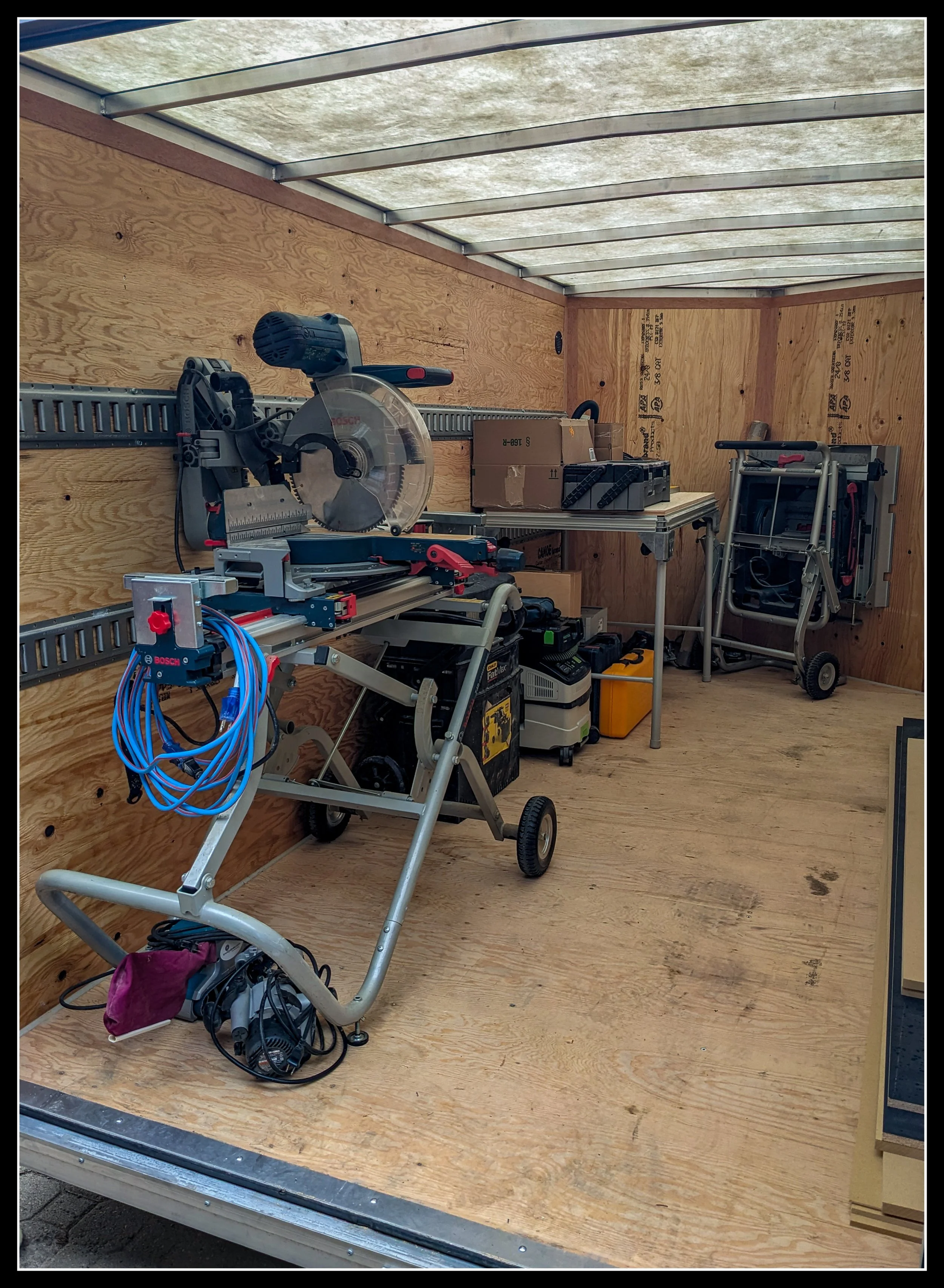 Mitre saw mounted on a portable stand inside a utility trailer, surrounded by jobsite tools, cords, and equipment.
