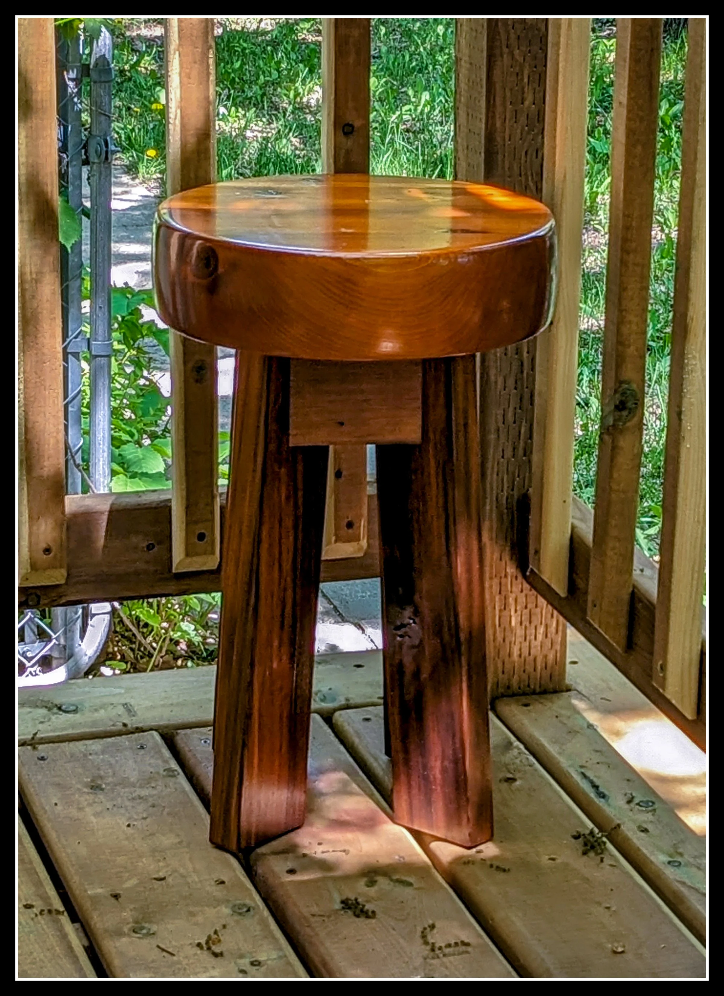 A wooden cedar stool placed on a wooden deck with a background of greenery, including plants and a fence.