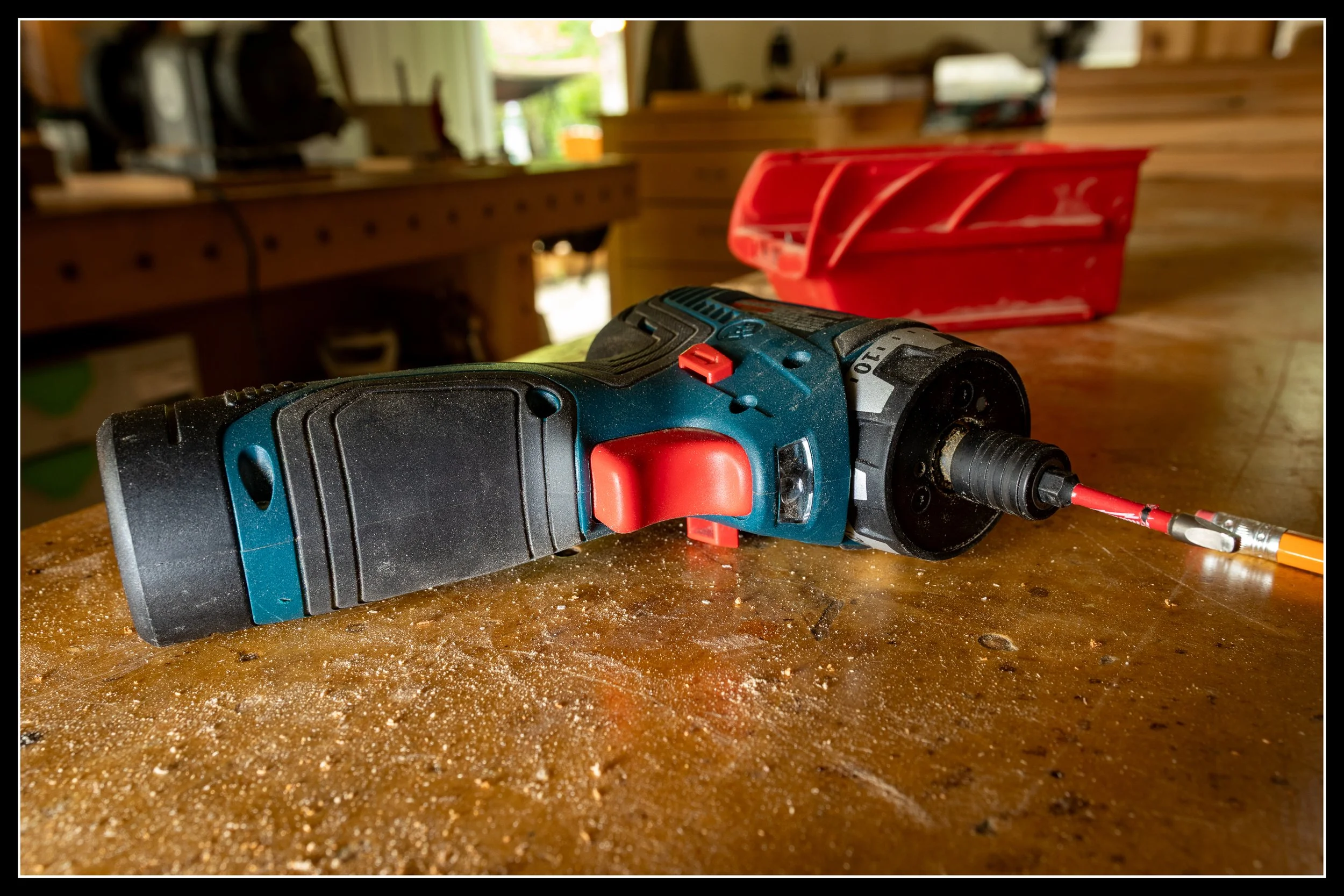 A compact 12V pocket driver resting on a wooden workbench, with a driver bit installed and other tools blurred in the background of a busy workshop.