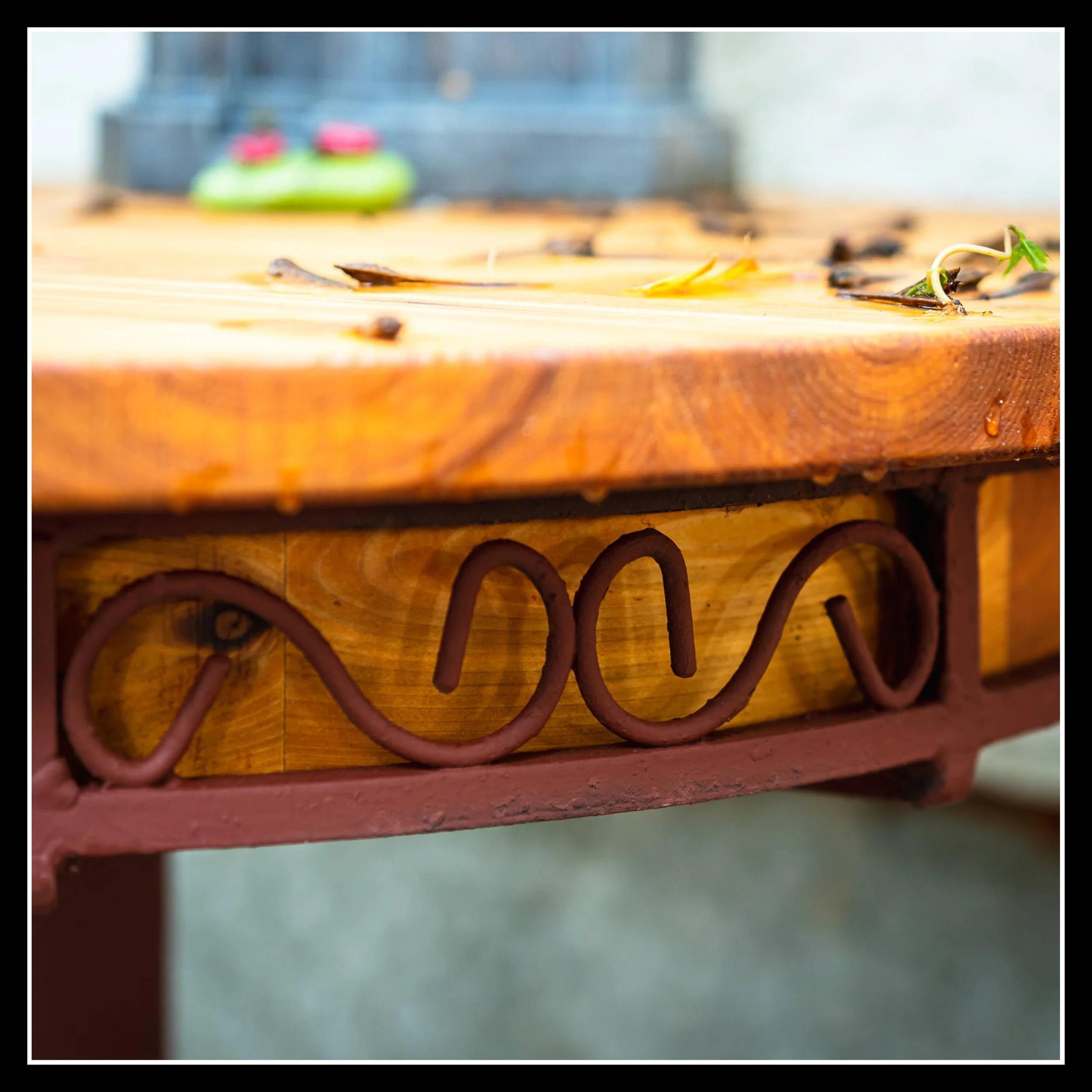 Close-up of a wooden cedar table with fallen leaves and a small green plant sprout, featuring decorative ironwork below the table surface.