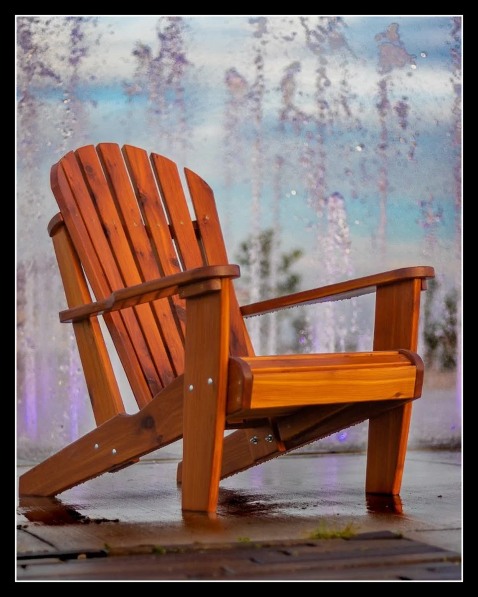 A wooden Adirondack chair on a wet deck with a blurred water fountain background.