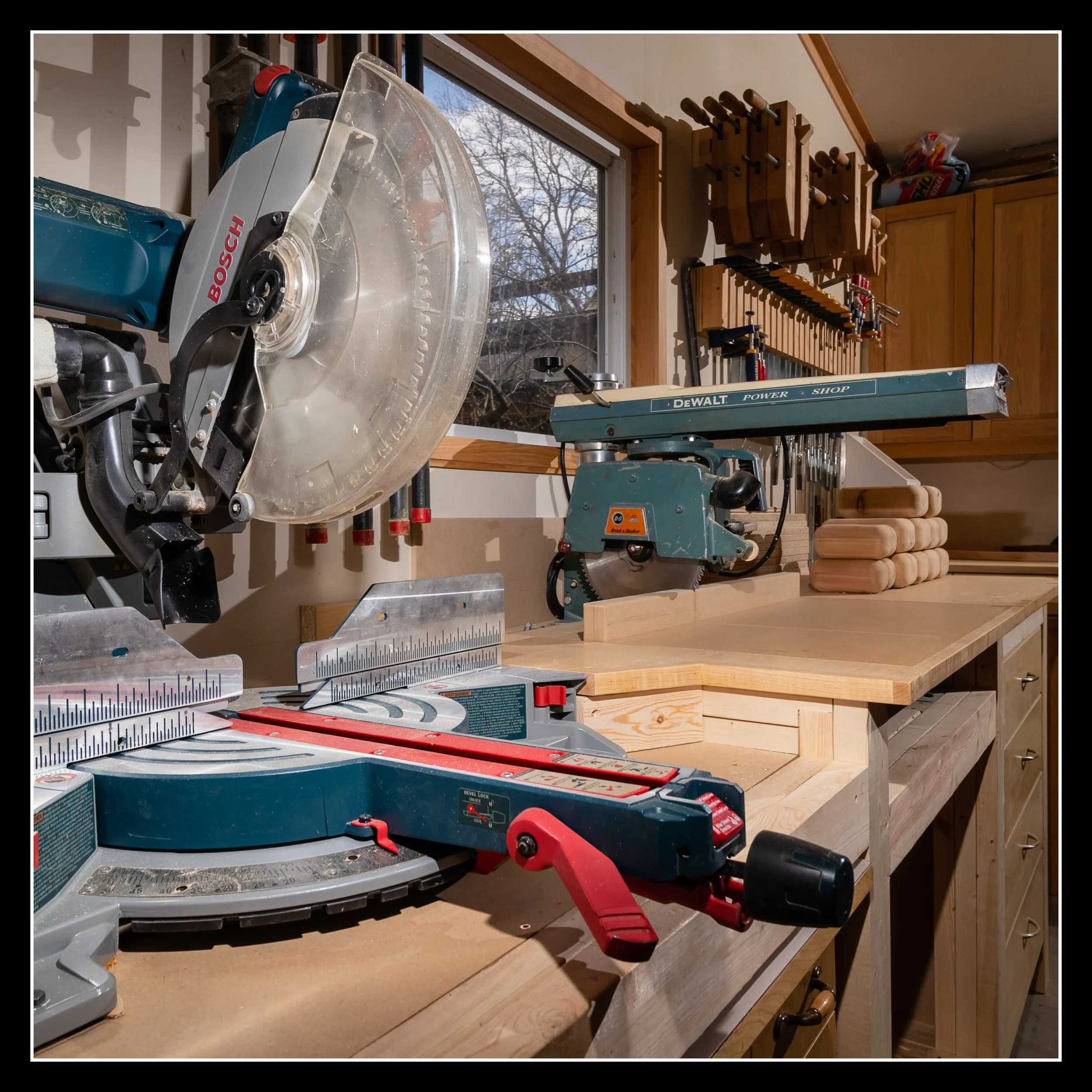 Bosch Axial‑Glide 12" mitre saw set up in a woodworking shop beside a radial arm saw, surrounded by cabinets, tools, and stacked lumber.