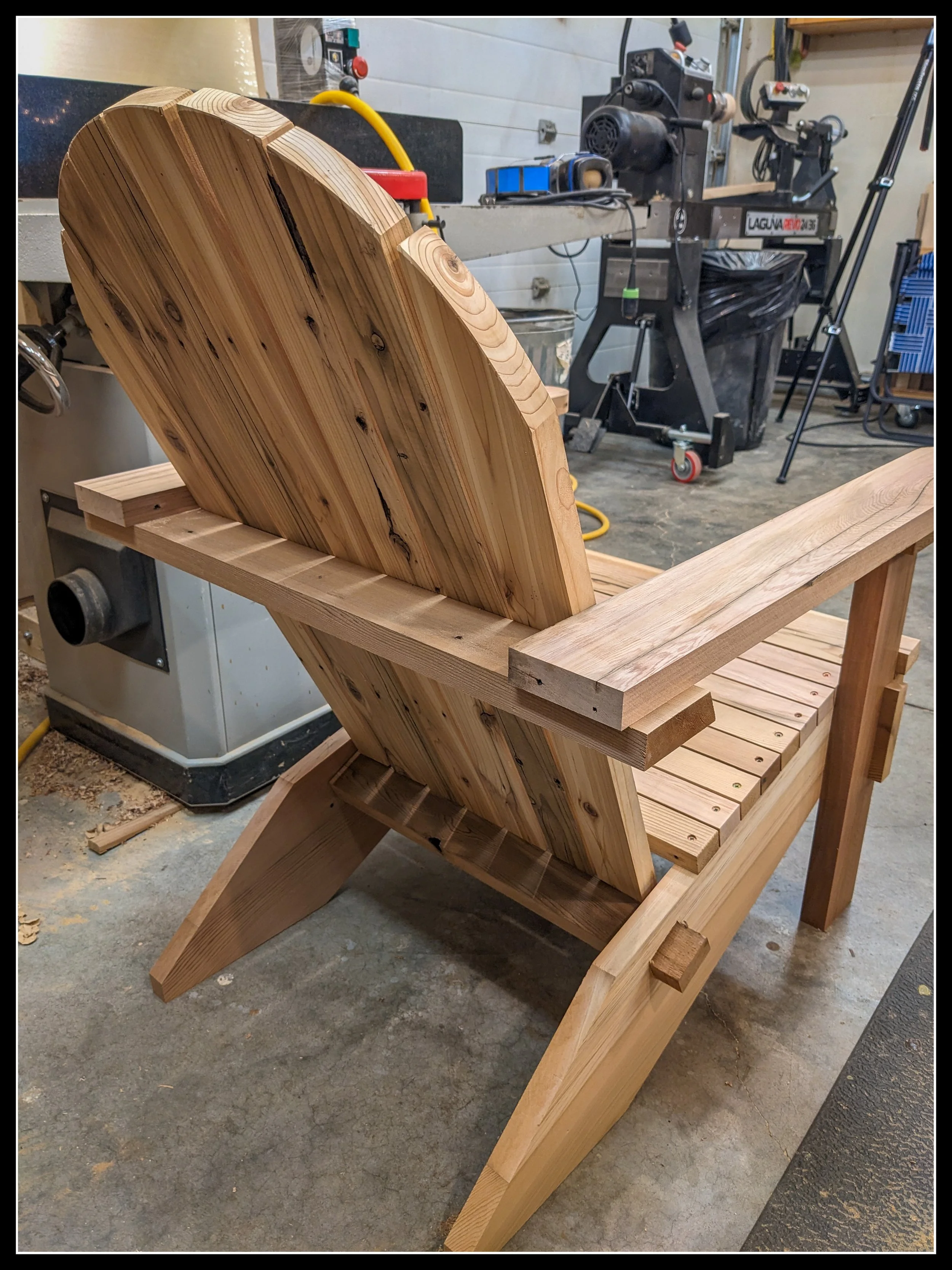 Partially built wooden Adirondack chair in a woodworking shop, fully assembled and ready for sanding and finish, with curved slat back, wide armrests, and tools visible in the background.