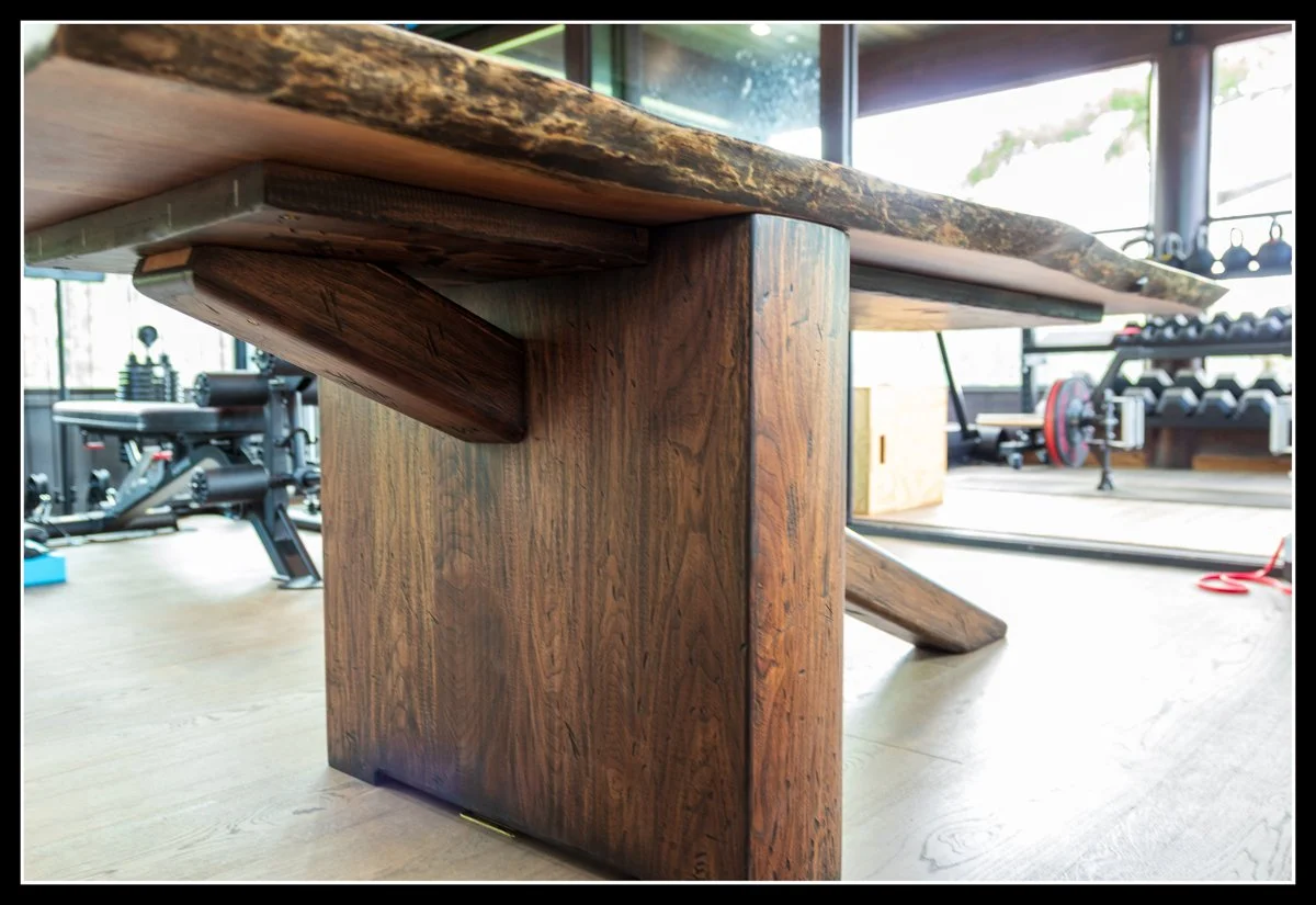 Close-up view of a large wooden conference table with a sturdy wooden leg, in a well-lit gym or fitness studio with exercise equipment and weights in the background.