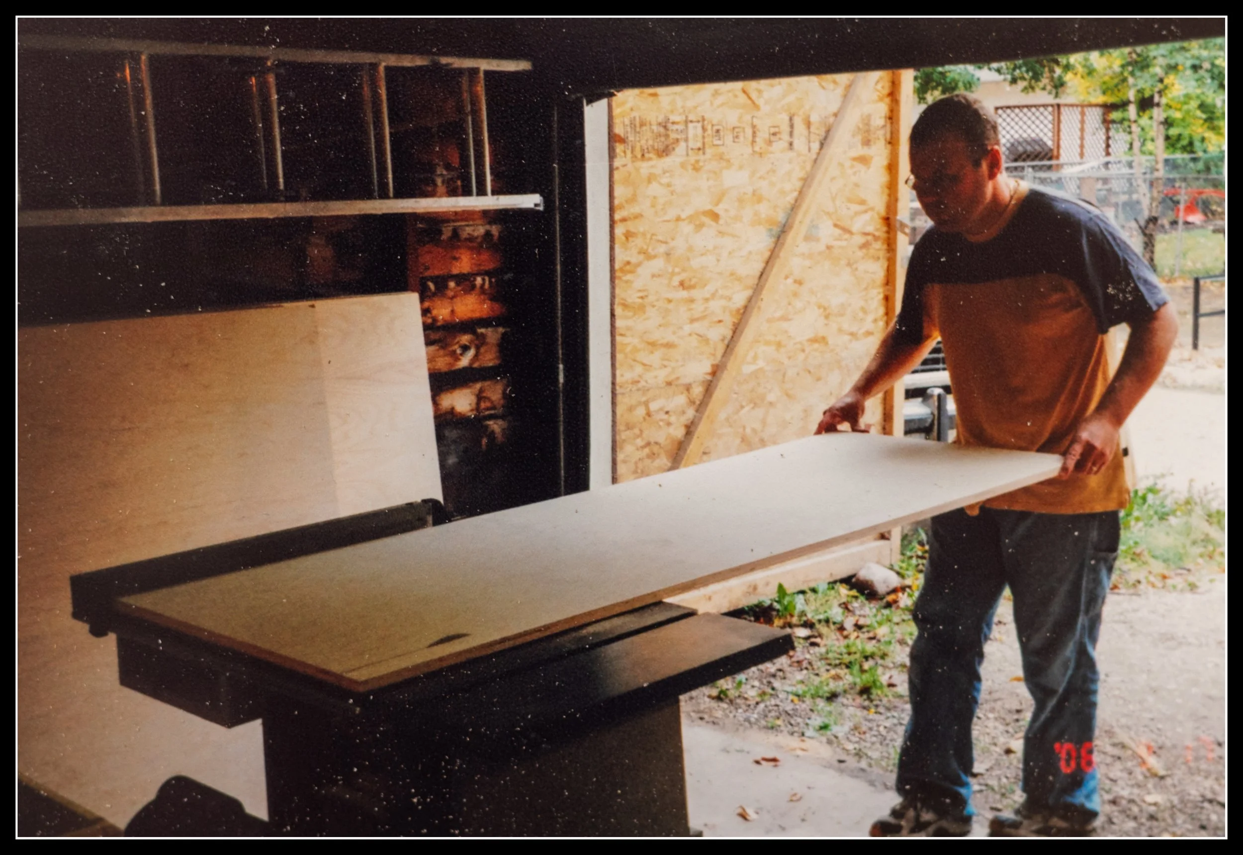 A young woodworker in his twenties guiding a large sheet of plywood across a Rockwell Beaver table saw in a garage workshop, with more sheets leaning against the wall and the garage door open to the yard.