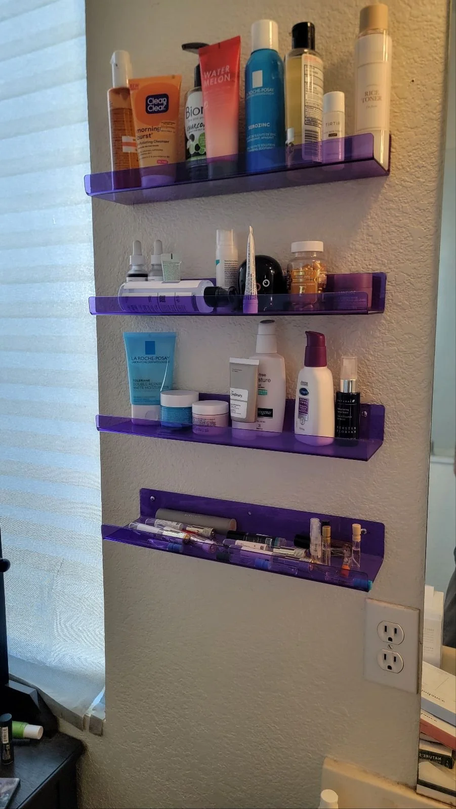 Mounted purple acrylic shelves on a wall holding various skincare, haircare, and cosmetic products. There's a power outlet below the shelves and a window with a light semi-transparent curtain next to it.