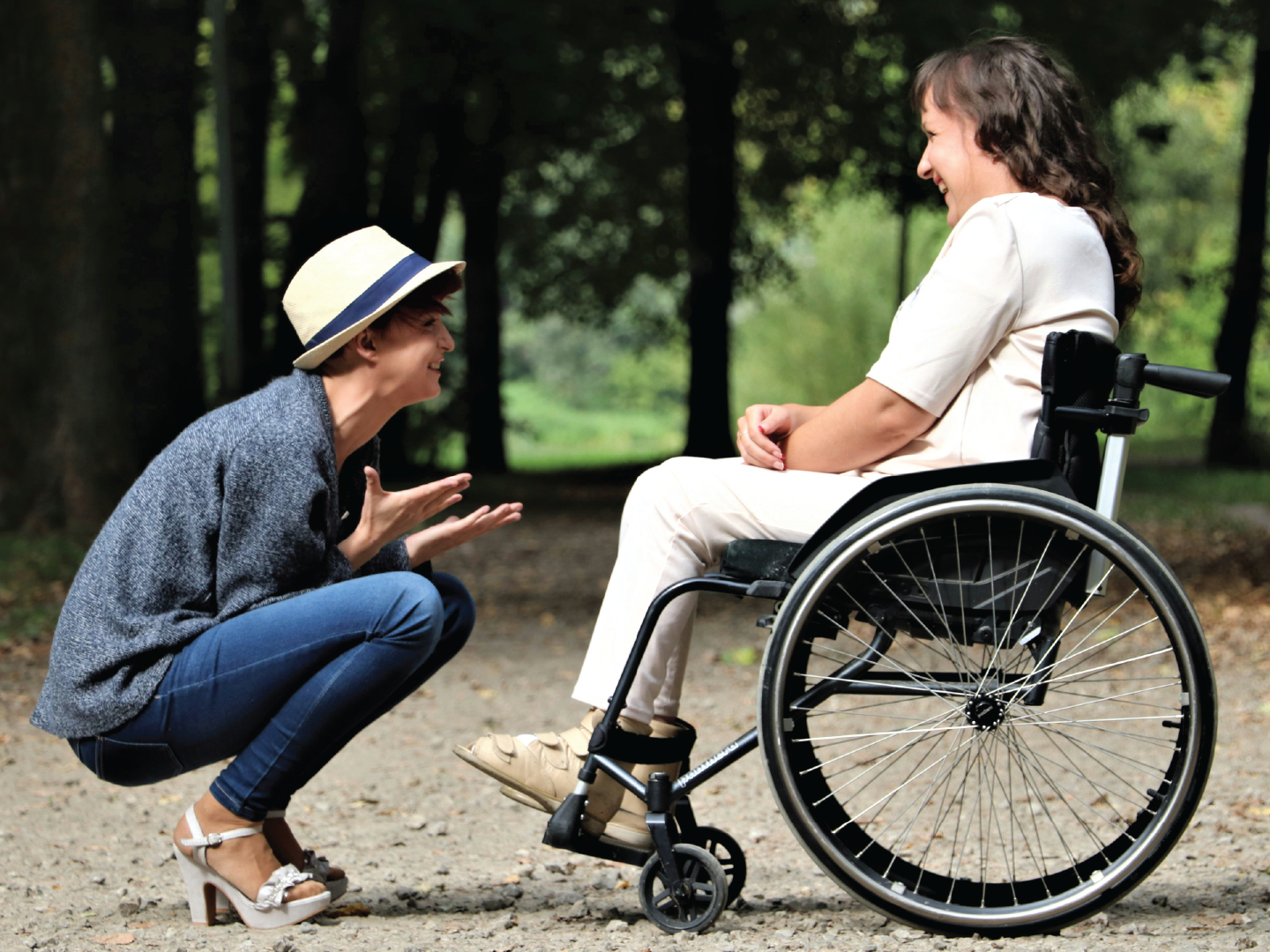 A woman in a wheelchair smiling and looking at a woman in casual clothes and a sun hat who is crouching and gesturing with her hands during an outdoor conversation in a forested area.