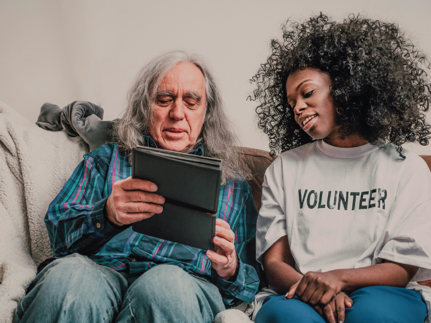 An elderly man with long gray hair and a young woman with curly hair sitting on a couch, looking at a photo album or book together. The woman is wearing a white T-shirt with the word 'VOLUNTEER' printed on it.