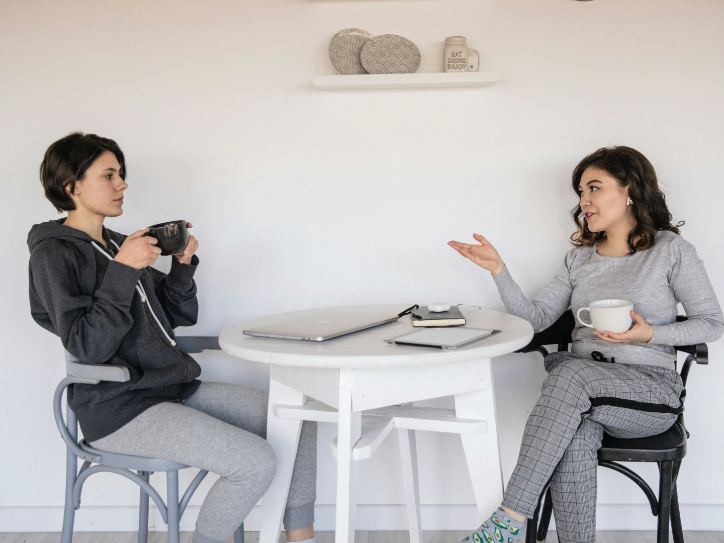 Two women sitting at a white table engaged in a conversation, each holding a cup and surrounded by laptops, in a minimalist room with a white wall and decorative items on a shelf.