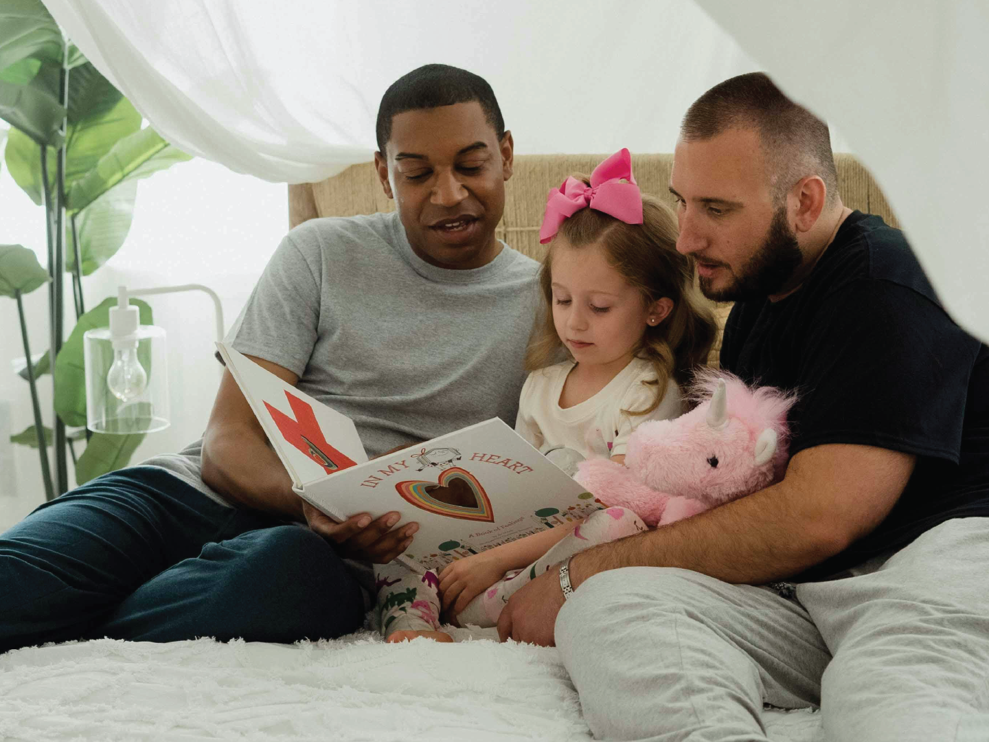 Family sitting on bed reading a children's book, with a young girl holding a pink stuffed unicorn, in a cozy bedroom with potted plants and sheer curtains.