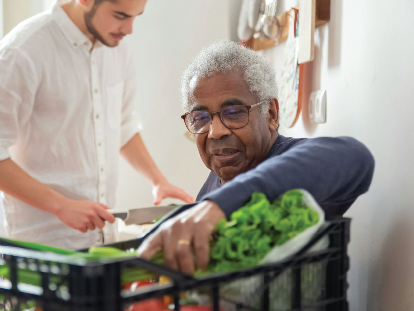 An elderly man with glasses sorting lettuce and vegetables in a crate while a younger man in a white shirt prepares food in the background.