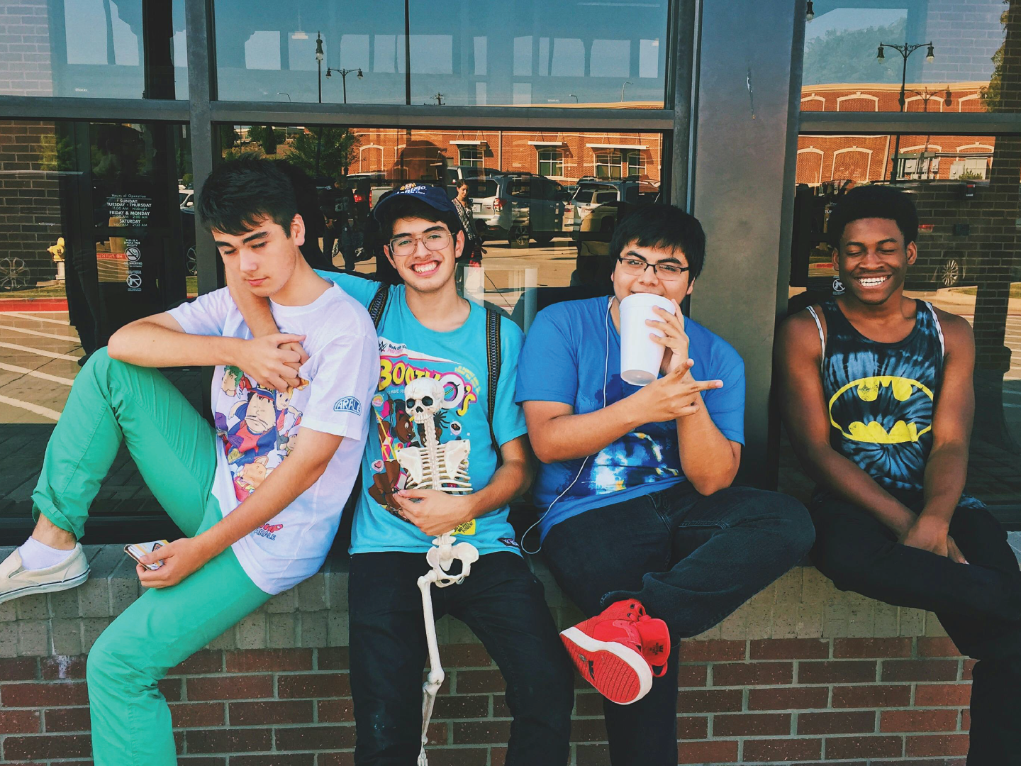 Four young men sitting outside a building on a brick ledge, smiling and wearing casual clothing, with a glass window behind them reflecting parked cars and buildings.