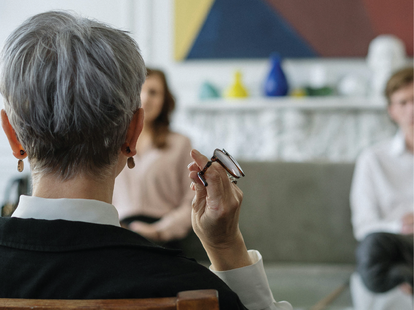 Older woman with short gray hair and earrings holding glasses while participating in a discussion in a room with abstract art and two blurred women in the background.