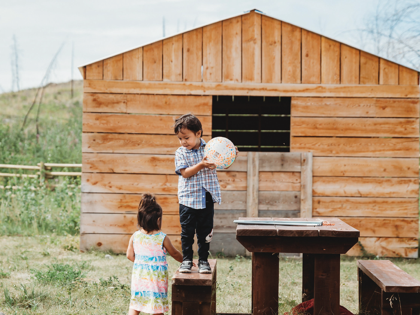 A boy and a girl playing outdoors in front of a wooden shed. The boy is holding a colorful ball while standing on a wooden platform, and the girl is reaching up to him.