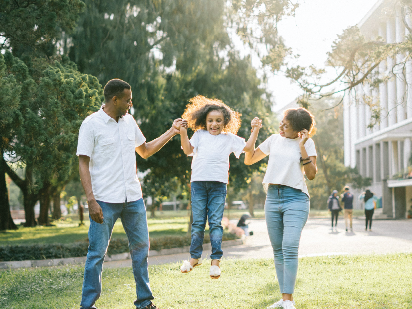 Family enjoying outdoors in park, parents swinging daughter between them, sunlight filtering through trees.