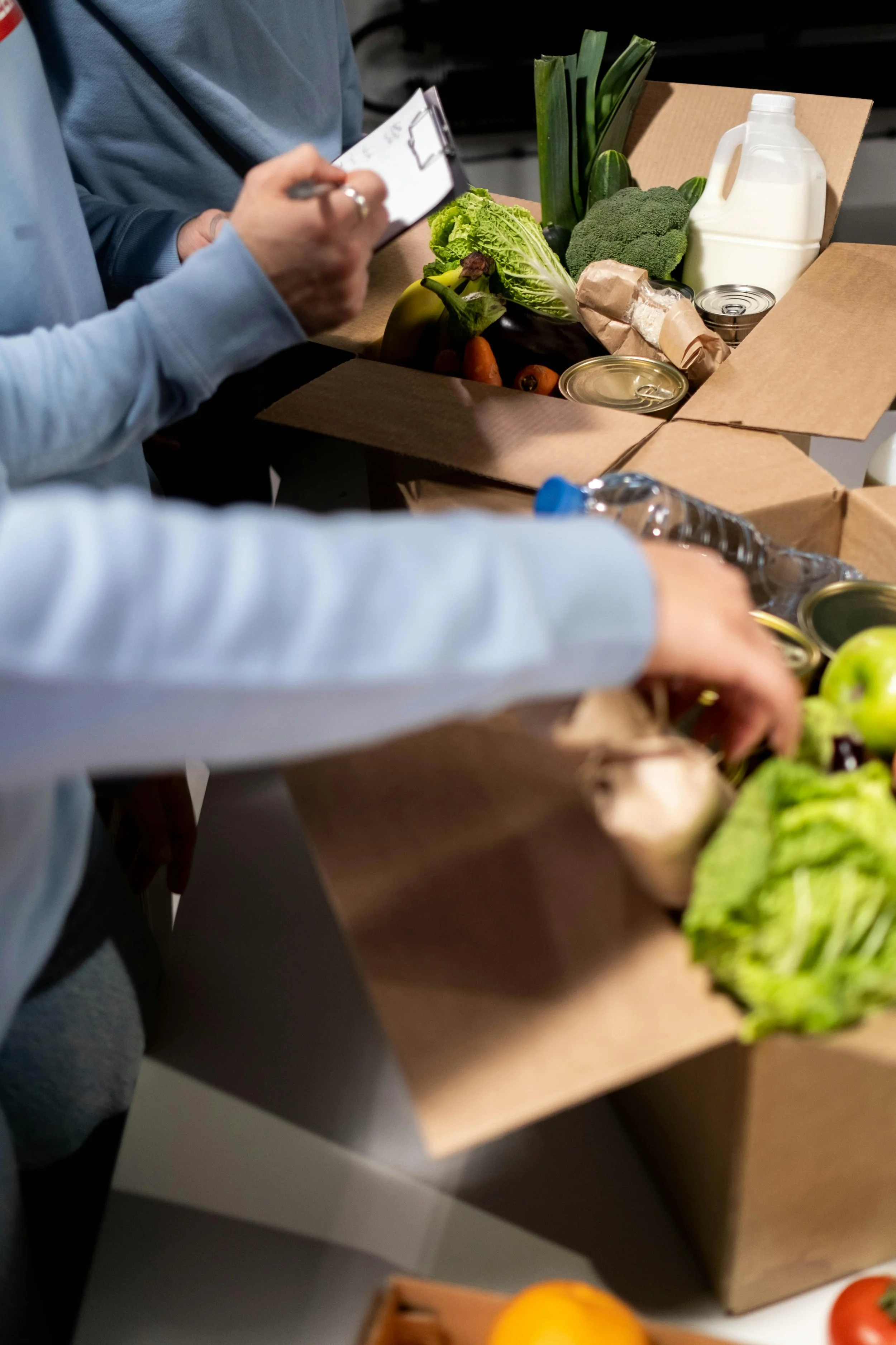 People packing a cardboard box with fresh vegetables and canned goods.