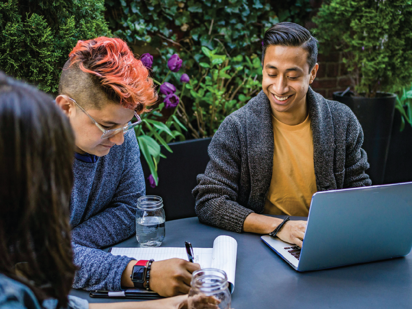 A group of young adults sitting at a table outdoors, engaged in discussion with one person using a laptop and another taking notes with a pen. The background features greenery and purple flowers.