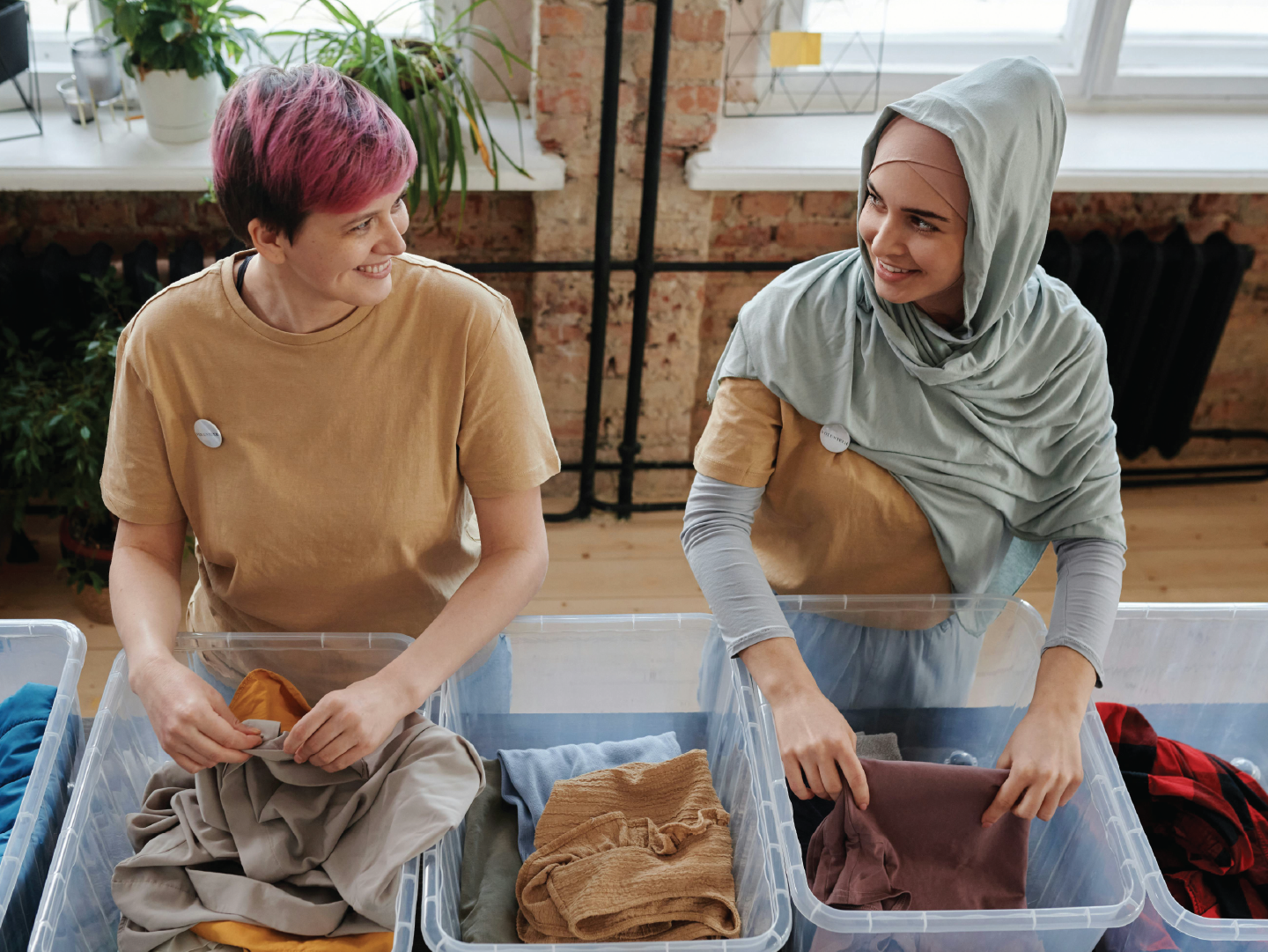 Two women sorting laundry in plastic bins, smiling and talking to each other in a bright, industrial-style room with large windows and plants.