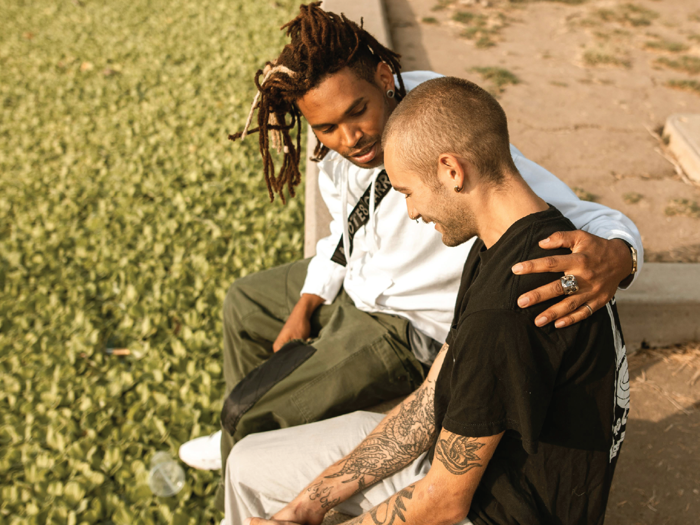 Two men sitting on a park bench sharing a moment of connection, one with long dreadlocks and the other with short hair, tattoos, and earrings.