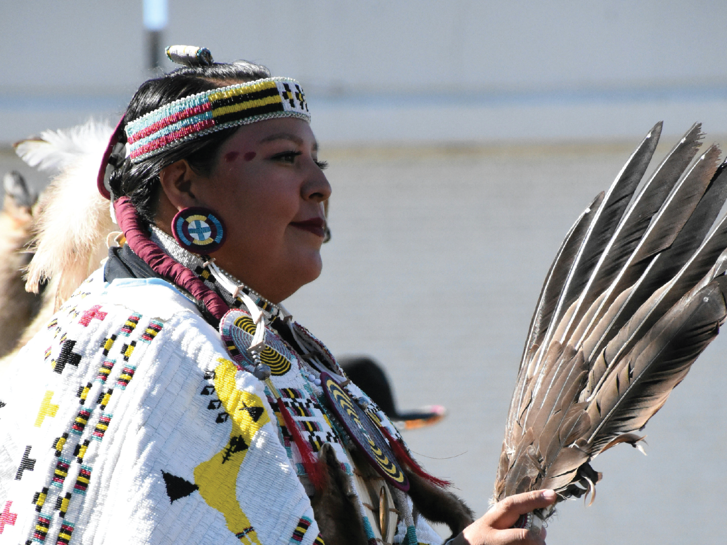 A Native American woman dressed in traditional attire with beaded jewelry, holding a fan of feathers against a body of water.