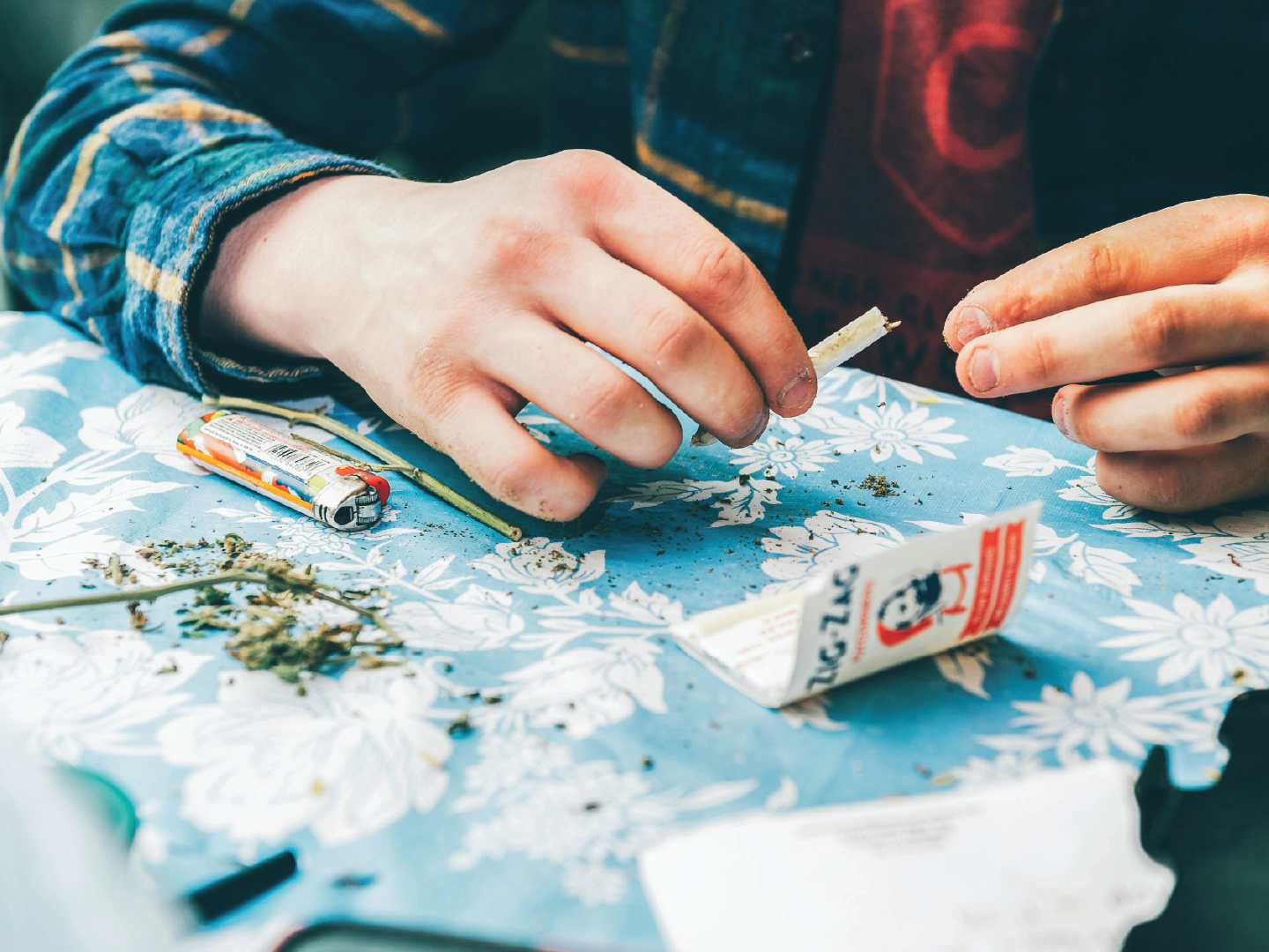 Close-up of a person rolling a small joint with marijuana on a floral tablecloth, with rolling papers, a lighter, and scattered marijuana visible.