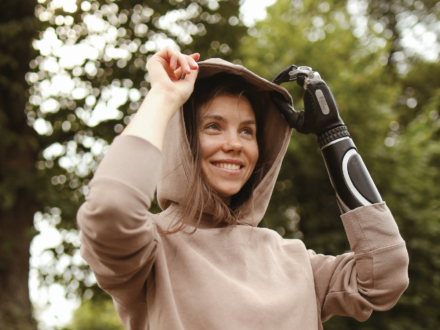 Young woman smiling outdoors, adjusting a hoodie with one hand and holding a prosthetic arm with a robotic hand with the other, surrounded by blurred green trees.