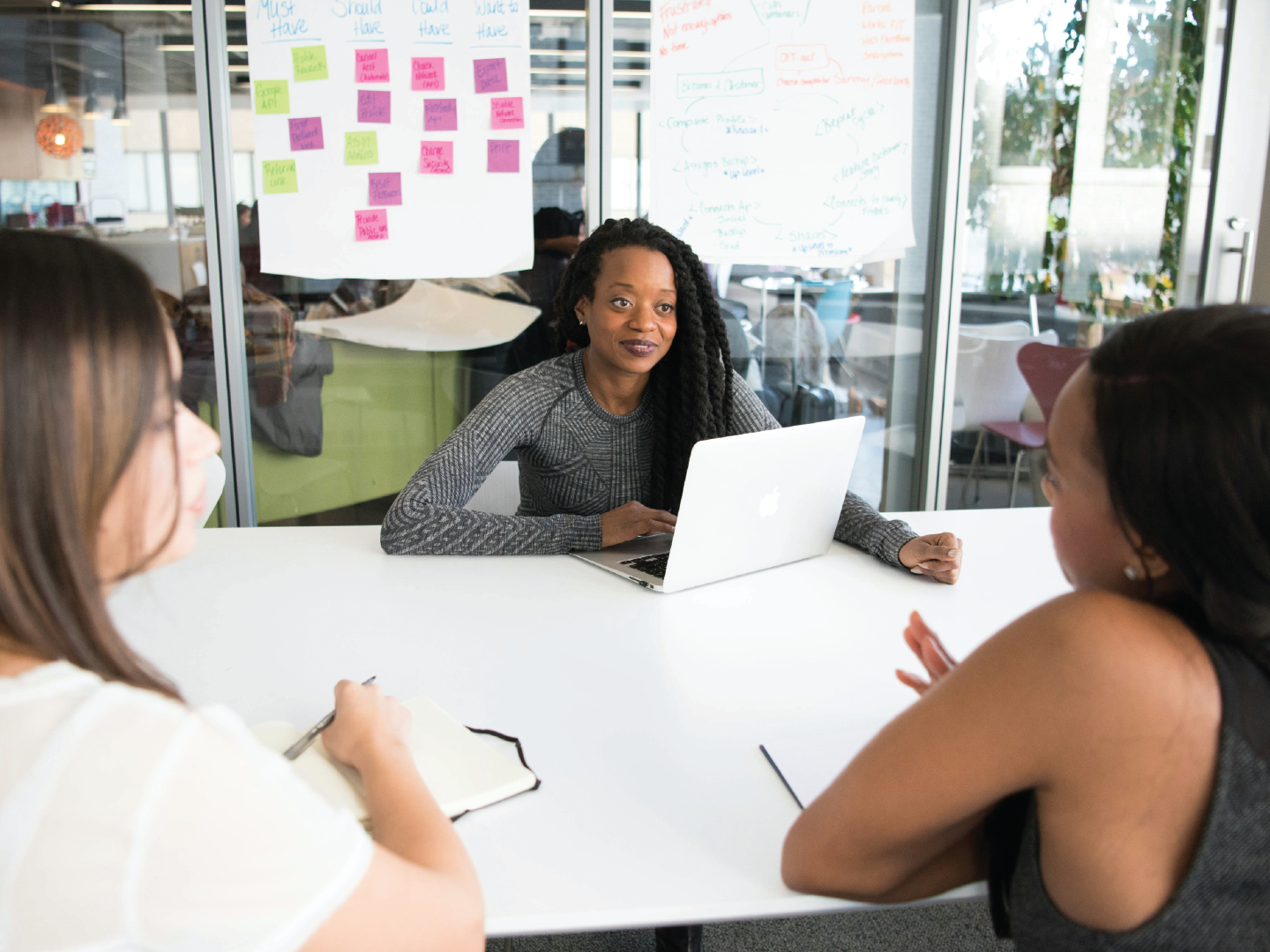 Three women in a meeting room, two are facing away and one is sitting at the end of a white table. The woman at the end has a laptop open in front of her. Behind her, there are large sheets of paper with notes and colorful sticky notes, and glass windows with a view of an office space and plants outside.