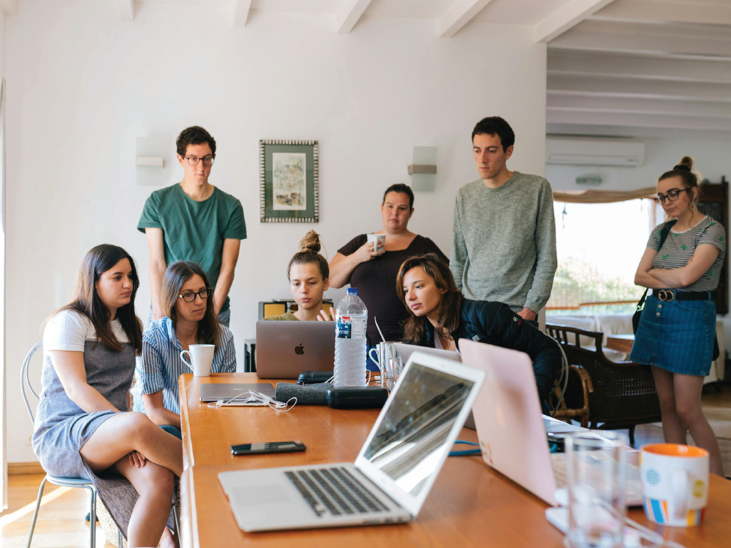 Group of seven young adults gathered around a conference table in a bright room, focused on a laptop screen, with some holding cups, and others standing nearby.