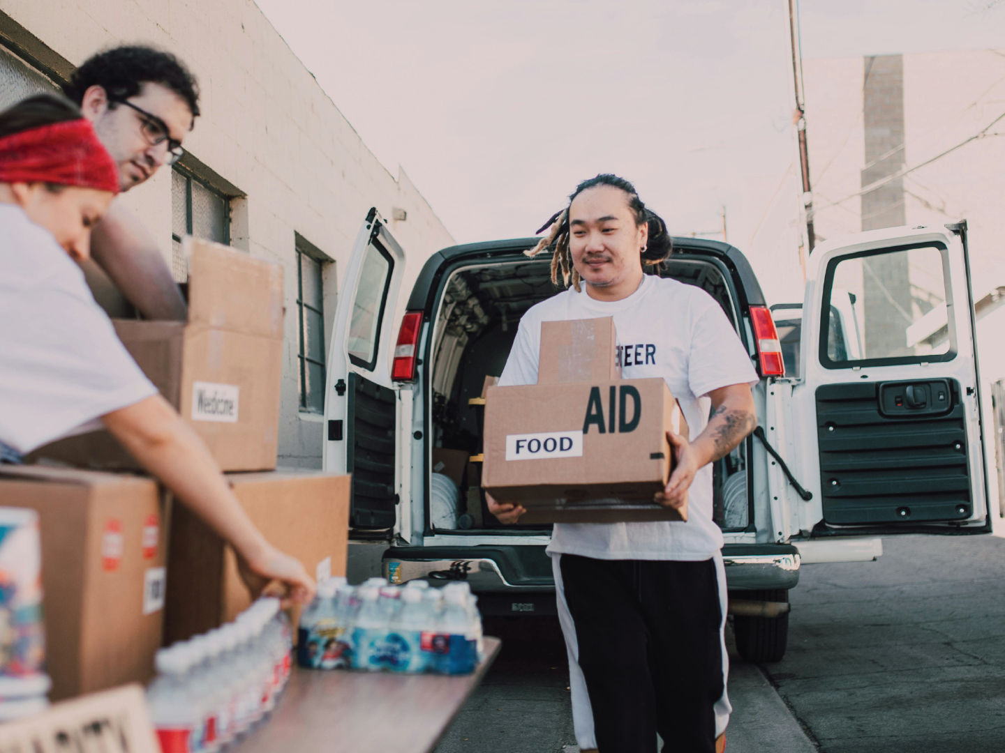 Man unloading boxes labeled 'Food Aid' from a van while two people prepare boxes of supplies, with a building and power lines in the background.