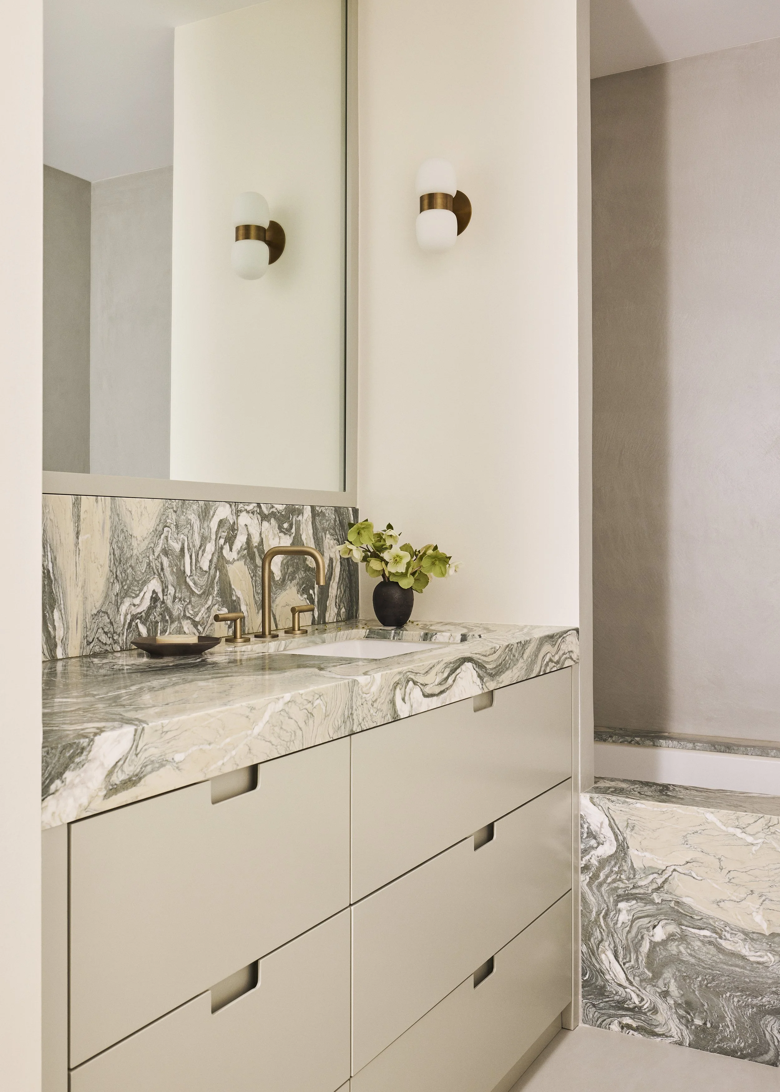 Modern bathroom vanity with a marble countertop, brass faucet, a small black vase with green flowers, a mirror, and wall-mounted light fixtures.