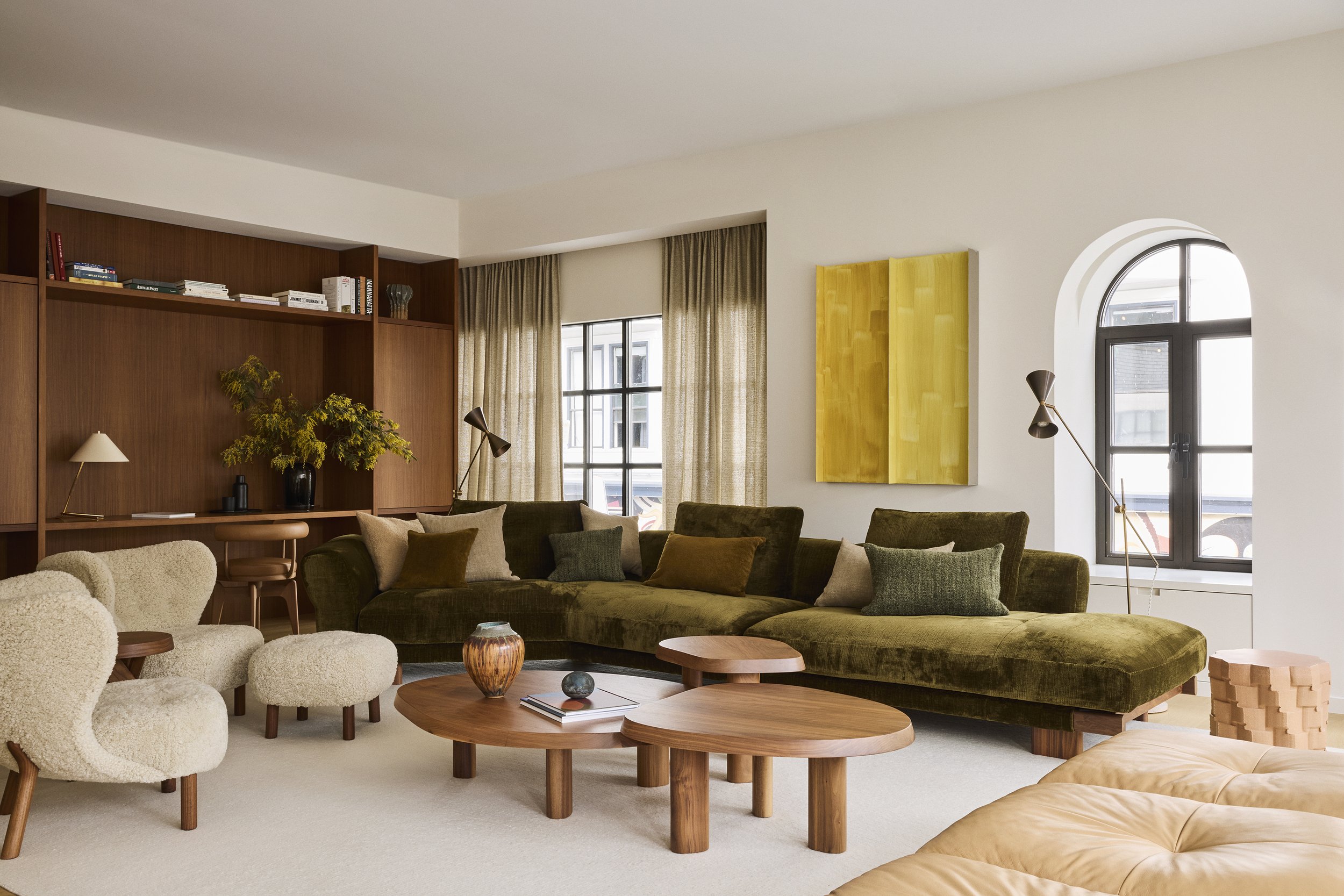 A living room with a large green sectional sofa, beige armchairs, wooden coffee tables, and a built-in wooden bookshelf with decor and books. There are floor lamps, a window with curtains, and a yellow abstract painting on the wall.