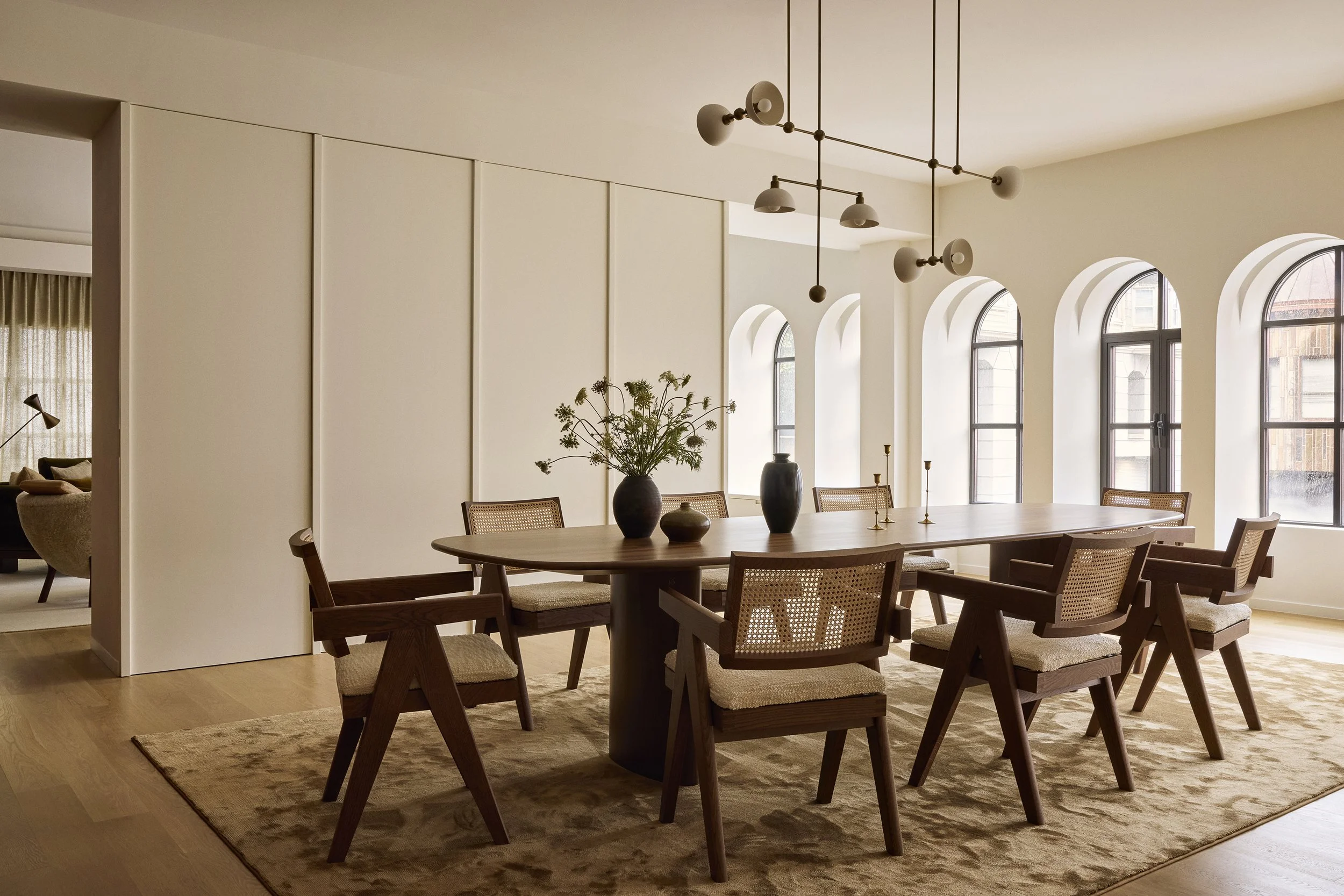 Modern dining room with wood table, eight chairs, vases, candlesticks, and pendant lighting, with large windows and light-colored walls.
