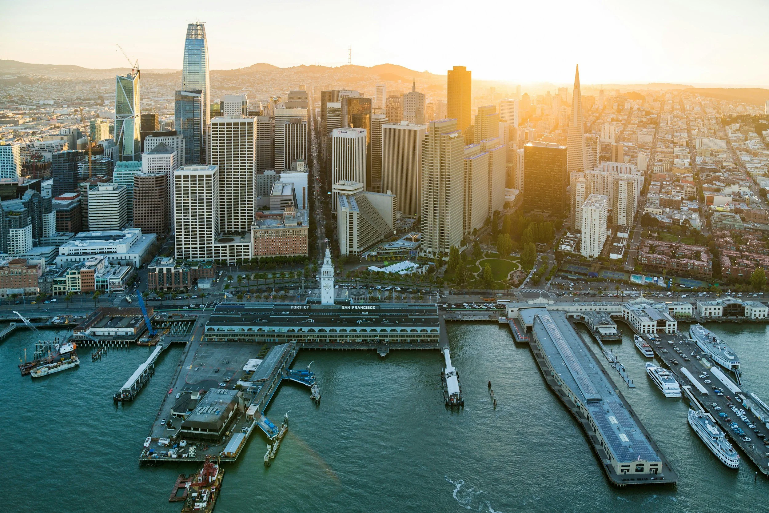 Aerial view of the San Francisco skyline at sunset, showing tall skyscrapers, the port with piers and boats, and a clock tower in the foreground.