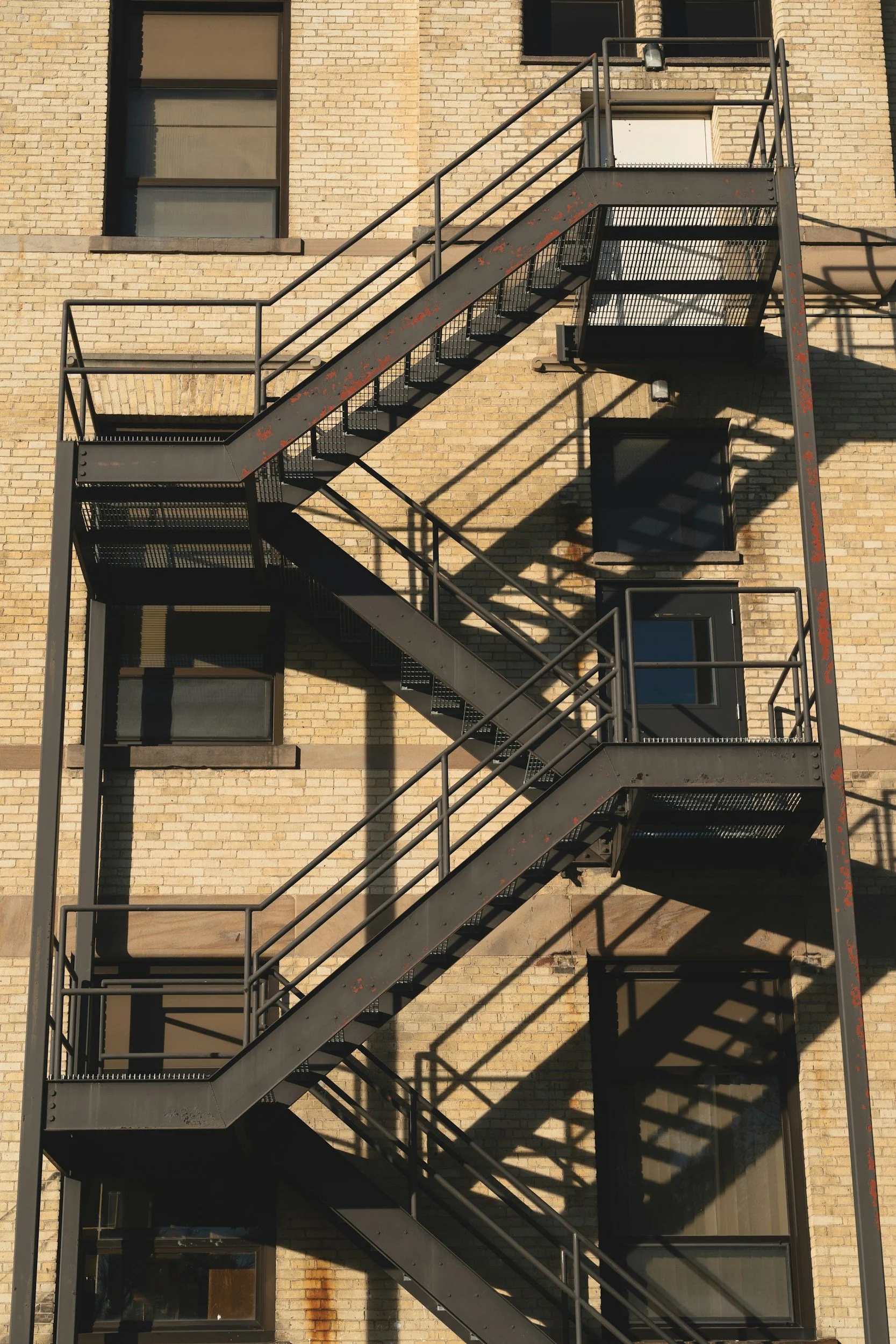 Metal fire escape staircase attached to a brick building with windows, casting shadows on the wall.