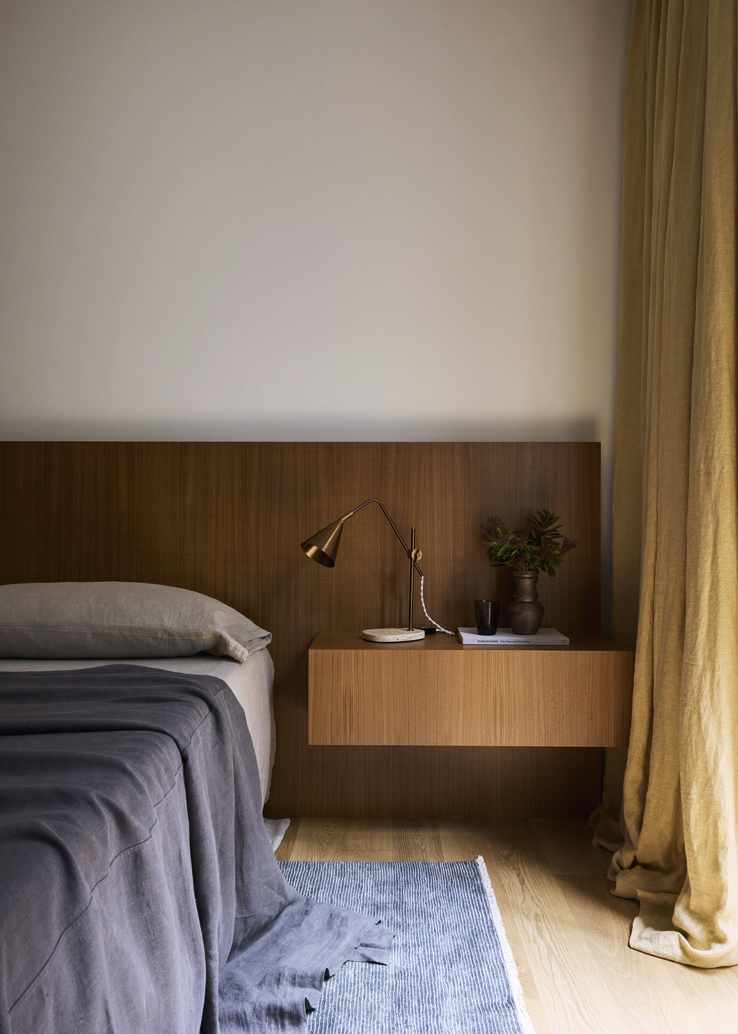 A bedroom with a wooden headboard, bedside table with a modern gold desk lamp, a vase with green plants, a small book, and a black cup. A gray bed with bedding and a striped blue and white rug on light wooden floor, with yellow curtains to the right.