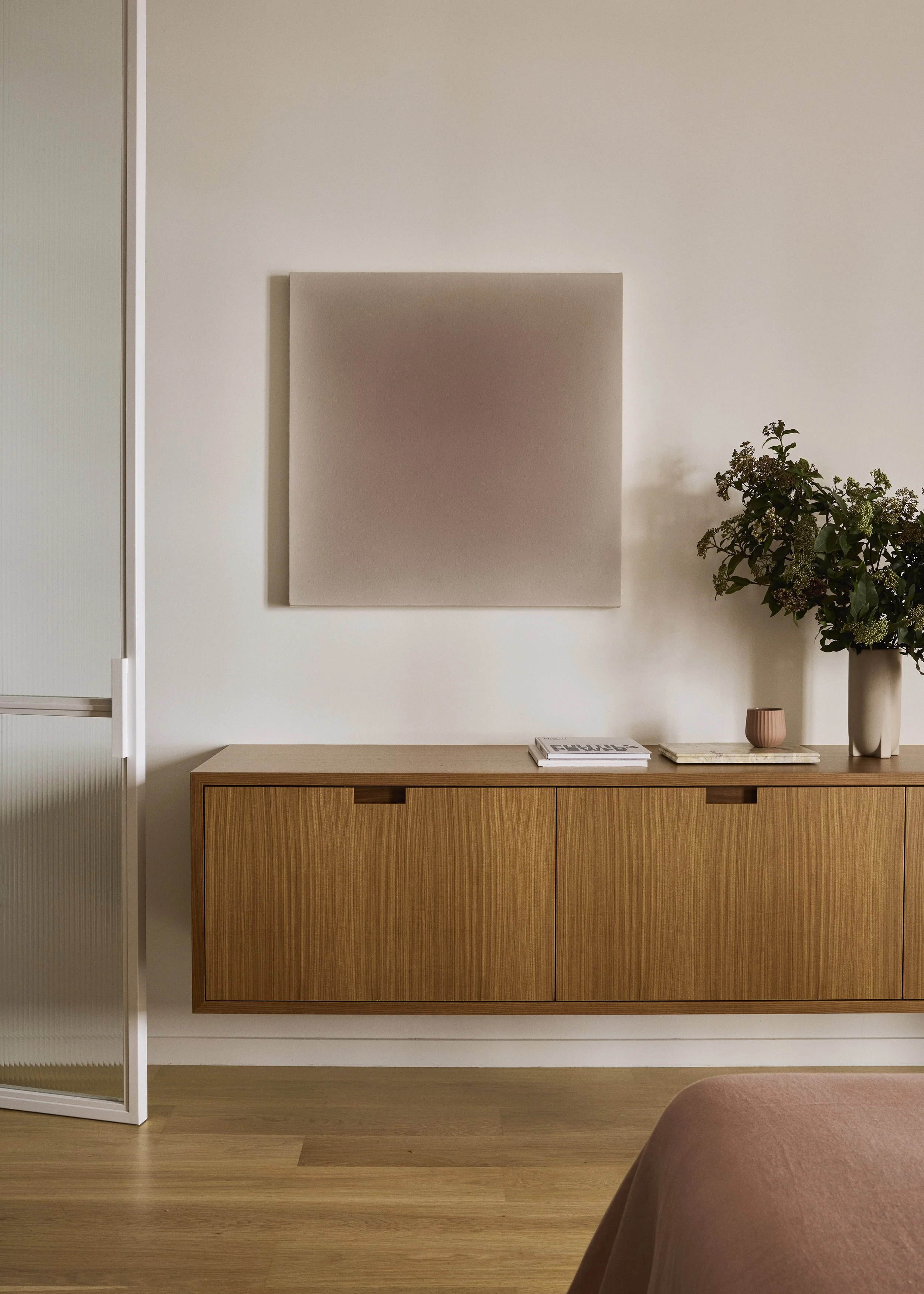 A minimalist room with a plain wall, a wooden sideboard, a small pink vase, a white flower arrangement, and a stack of magazines. There is a framed artwork on the wall above the sideboard.