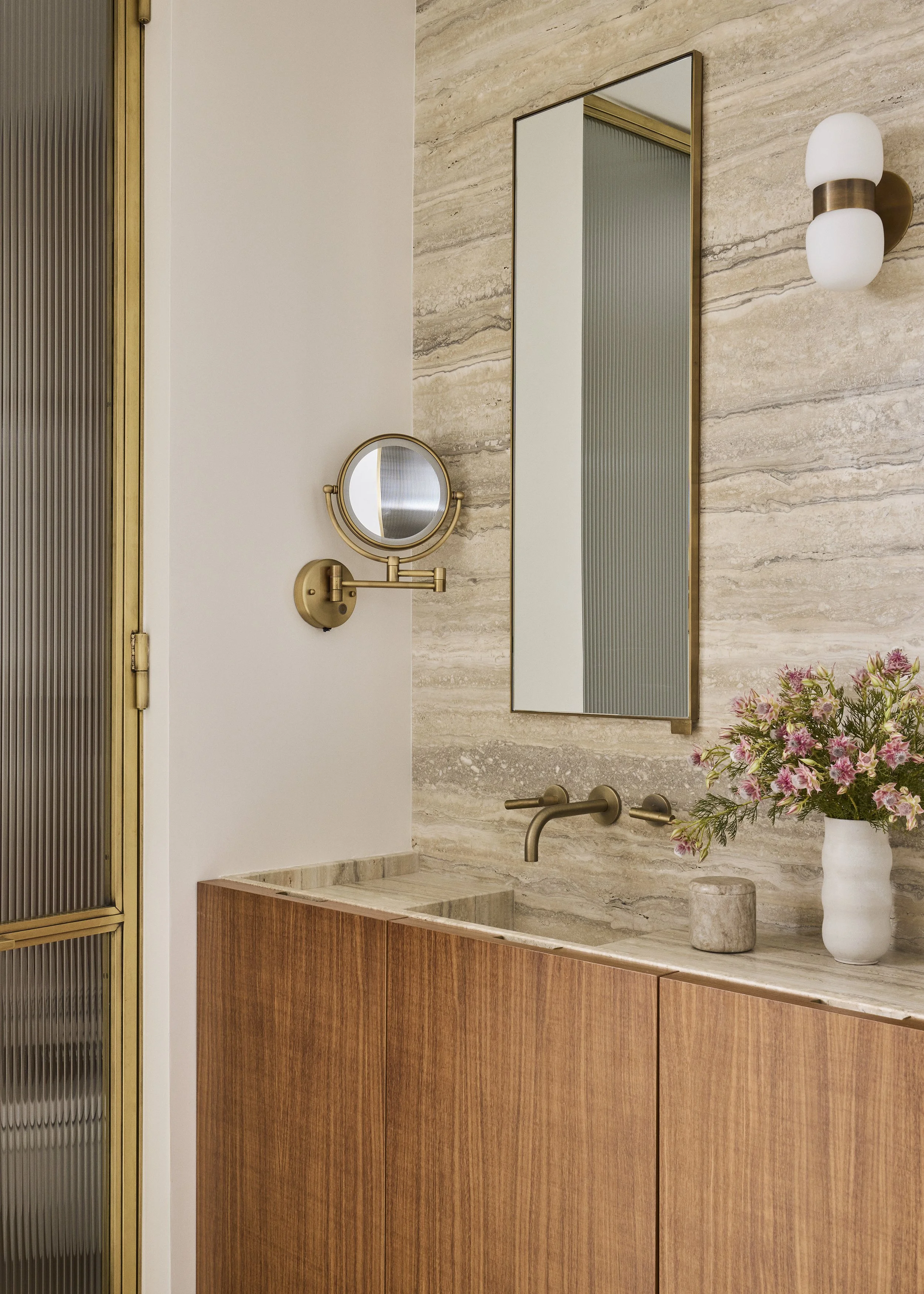 A modern bathroom vanity with wooden cabinetry, a marble countertop, and a wall-mounted faucet. There is a large mirror with a slim frame, a smaller round mirror attached to a wall-mounted magnifying mirror, a wall light fixture with two frosted glass globes, and a vase of pink flowers on the countertop.