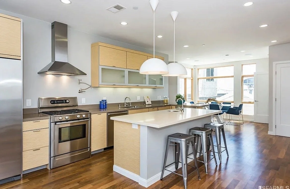 Modern open-concept kitchen and dining area with light wood cabinets, stainless steel appliances, a central island with stools, and a dining area with blue chairs near large windows allowing natural light.