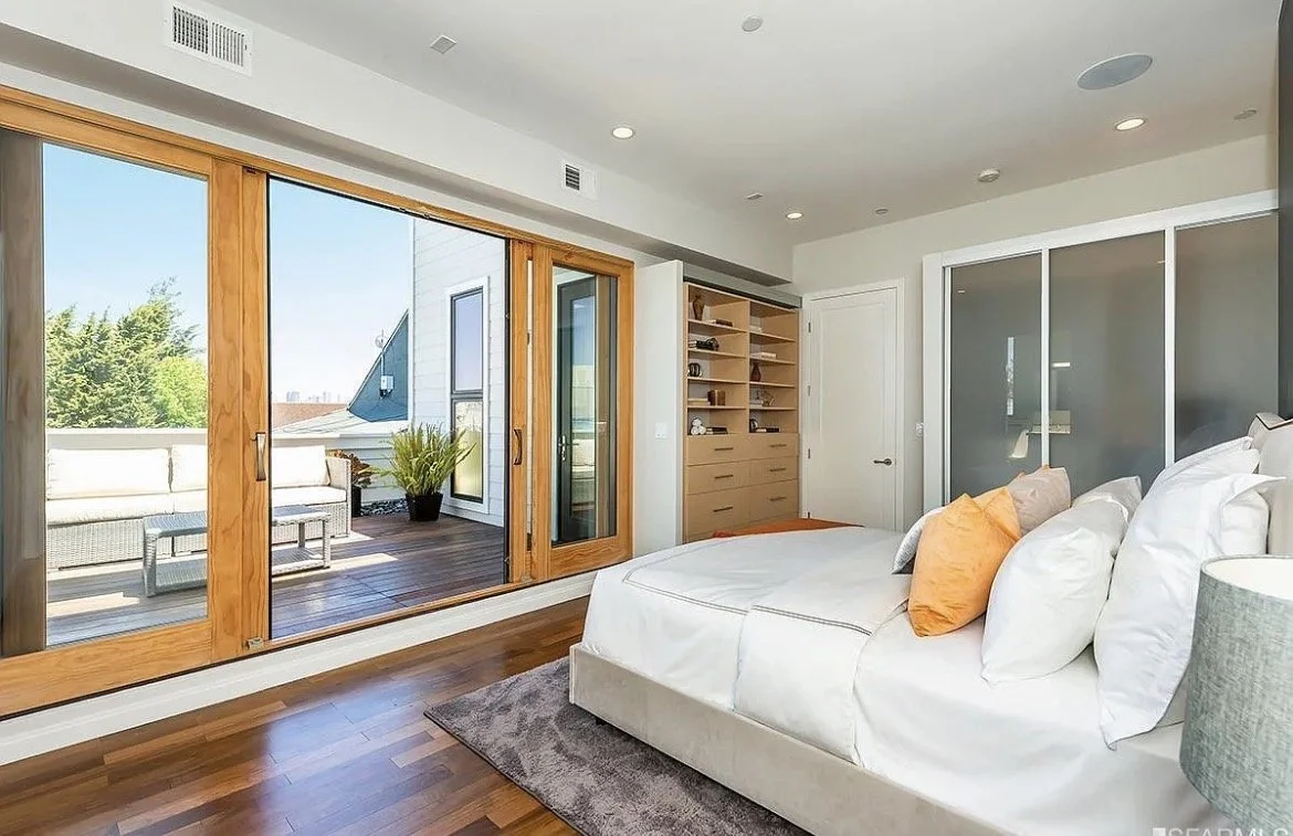Bedroom with large wooden sliding glass doors leading to a balcony with outdoor seating, white bed with orange pillows, wood flooring, and a built-in wooden shelf.
