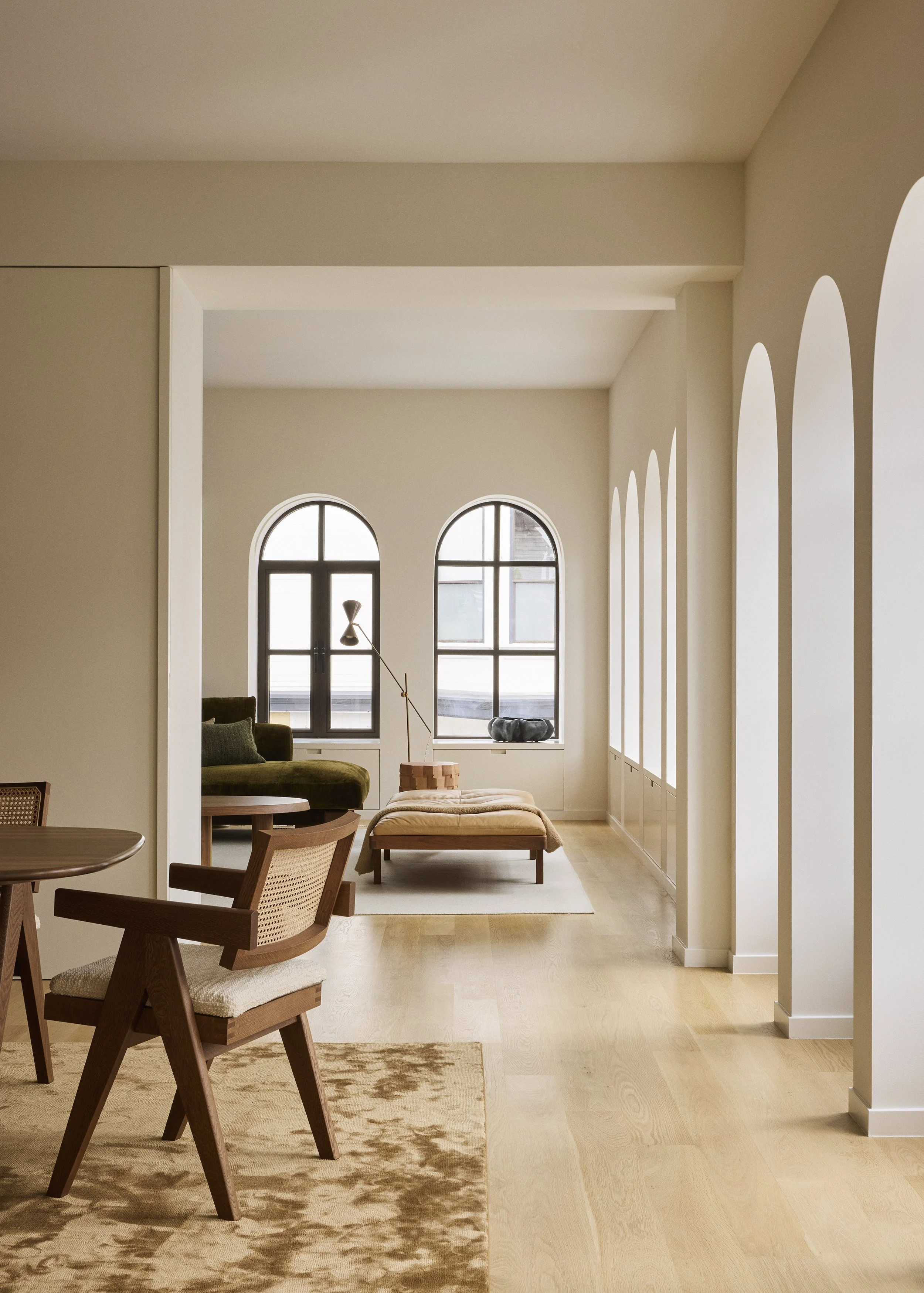 Modern living room with arched windows, a green sofa, a beige ottoman, a black sculpture, and sunlight streaming in.
