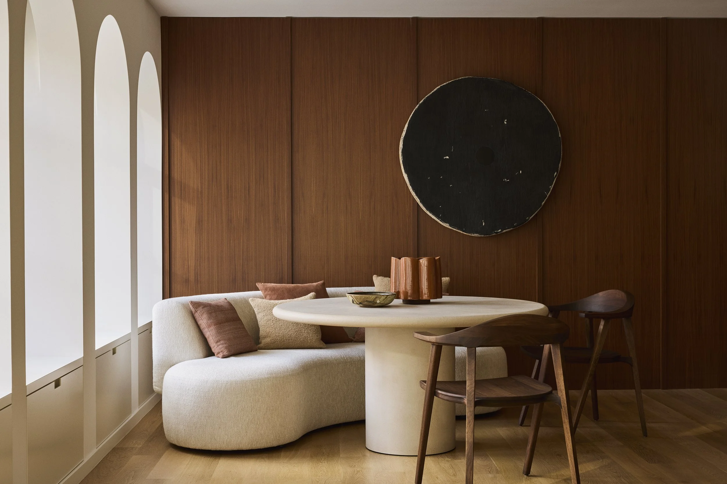 Contemporary dining area with a curved beige sofa, a round white table, and two wooden chairs, set against a wood-paneled wall with an abstract black circular artwork.