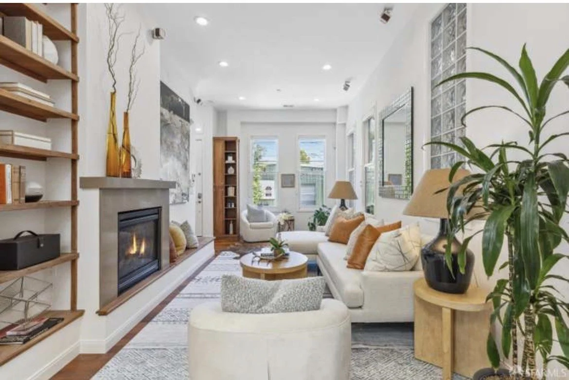 Bright living room with white walls, large windows, white sofas with orange pillows, a fireplace, wooden shelves, potted plants, and framed wall art.