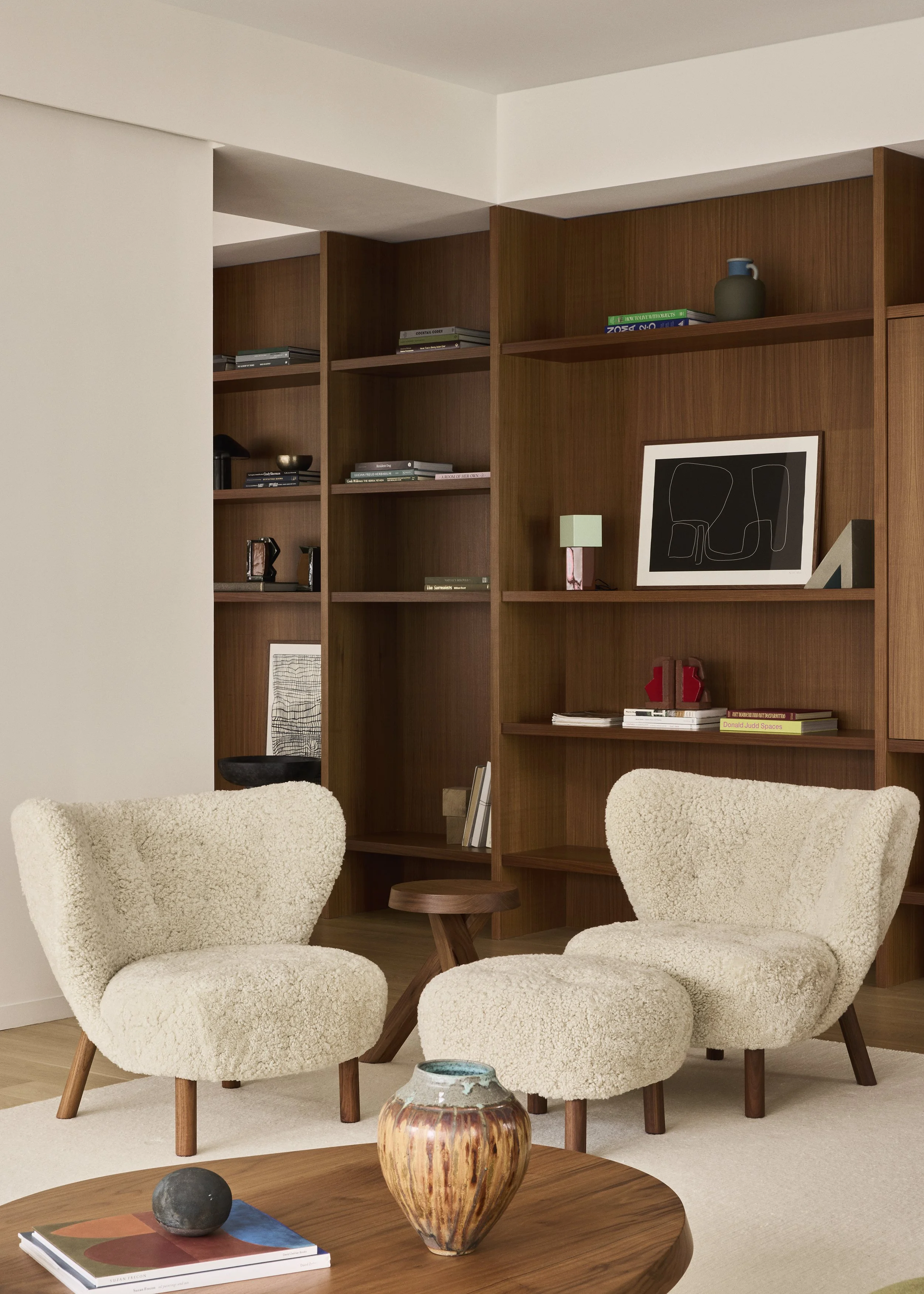A cozy living room corner with two cream-colored, plush armchairs with wooden legs. Between them is a small, round wooden side table. In front, there’s a wooden coffee table with a decorative vase, a round stone, and a book. Behind the chairs is a large wooden bookshelf with various books, framed art, and decorative objects.