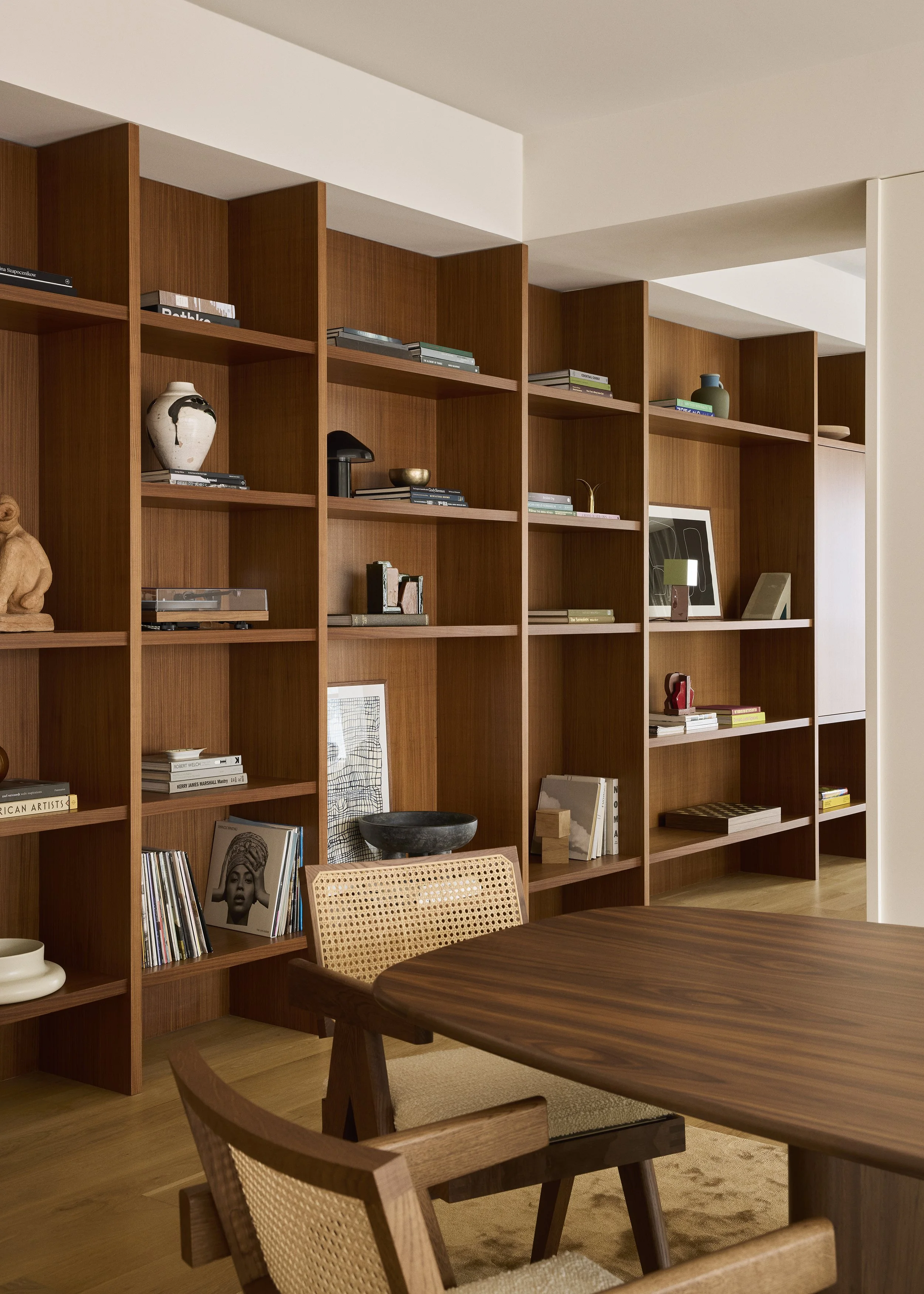 A wooden bookshelf filled with books, vases, and artwork in a modern living room with a wooden dining table and chairs.