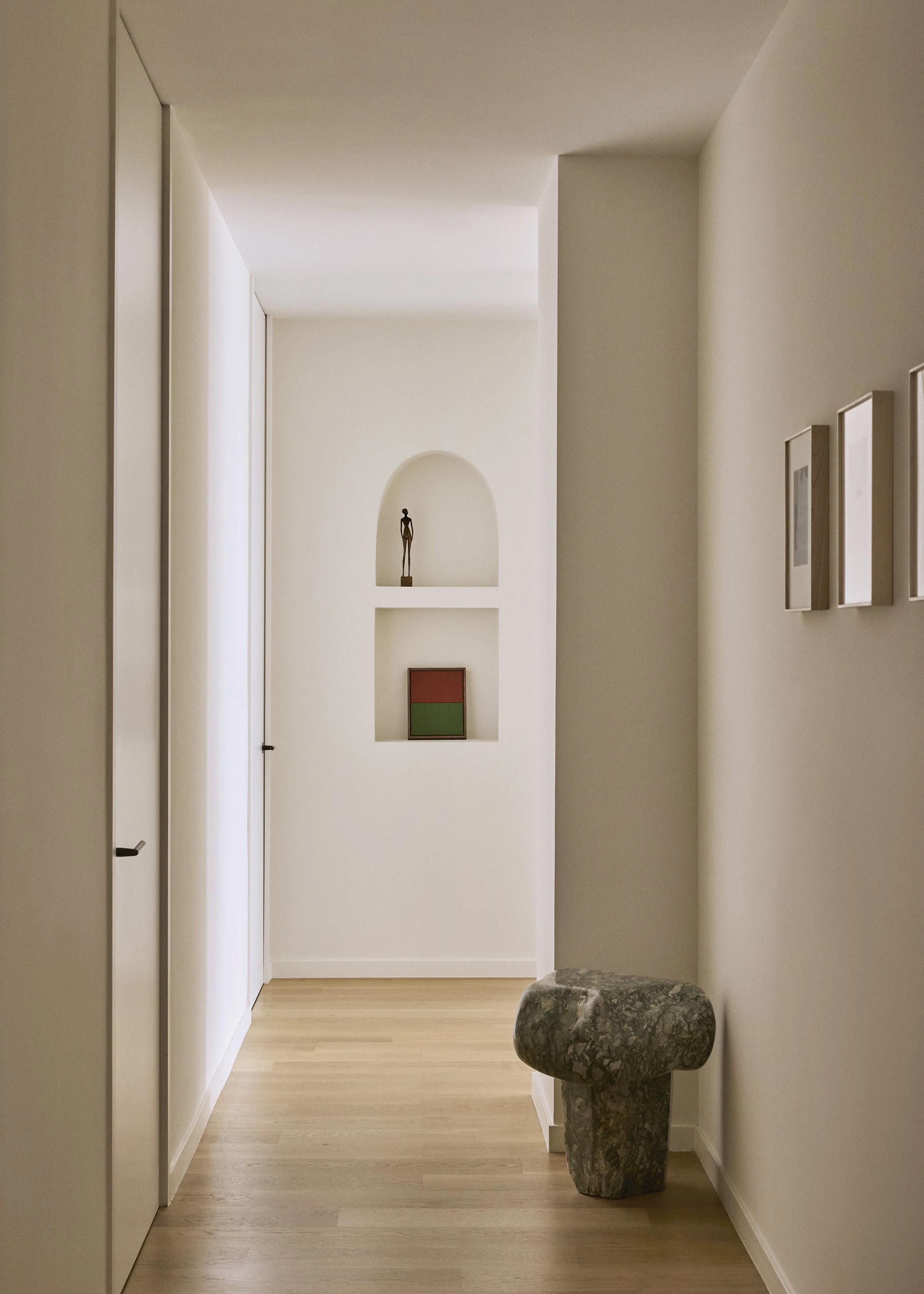A minimalist hallway with white walls, light wood flooring, and built-in shelves with decorative objects, including a small sculpture, framed artwork, and small pictures.
