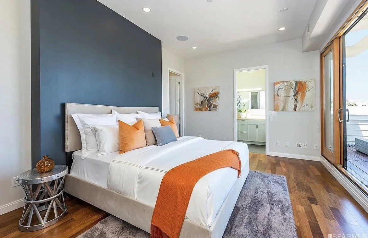 Modern bedroom with a large bed, orange and gray throw pillows, and a matching orange blanket. The room has wooden flooring, abstract artwork on the walls, a sliding glass door, and a small bedside table with a decorative item.
