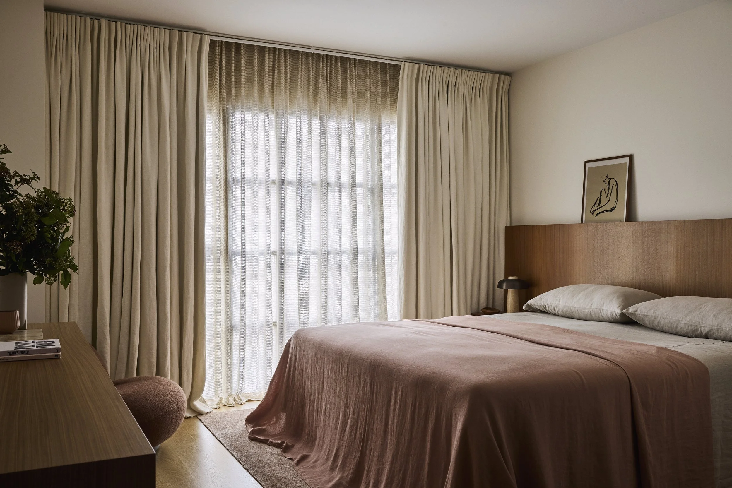 A cozy hotel bedroom featuring a large bed with white pillows and a peach-colored bedspread, a wooden headboard, a bedside lamp, a framed line art sketch on the wall, a dresser with books and a potted plant, and beige curtains covering a large window.
